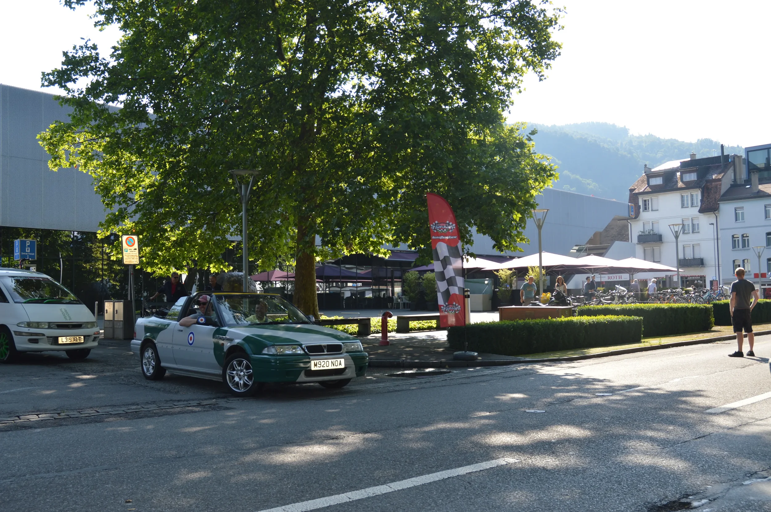 Street scene with cars parked near a large tree, a graffiti-covered flag, and an outdoor seating area with umbrellas. People are walking and sitting, with buildings and mountains in the background.