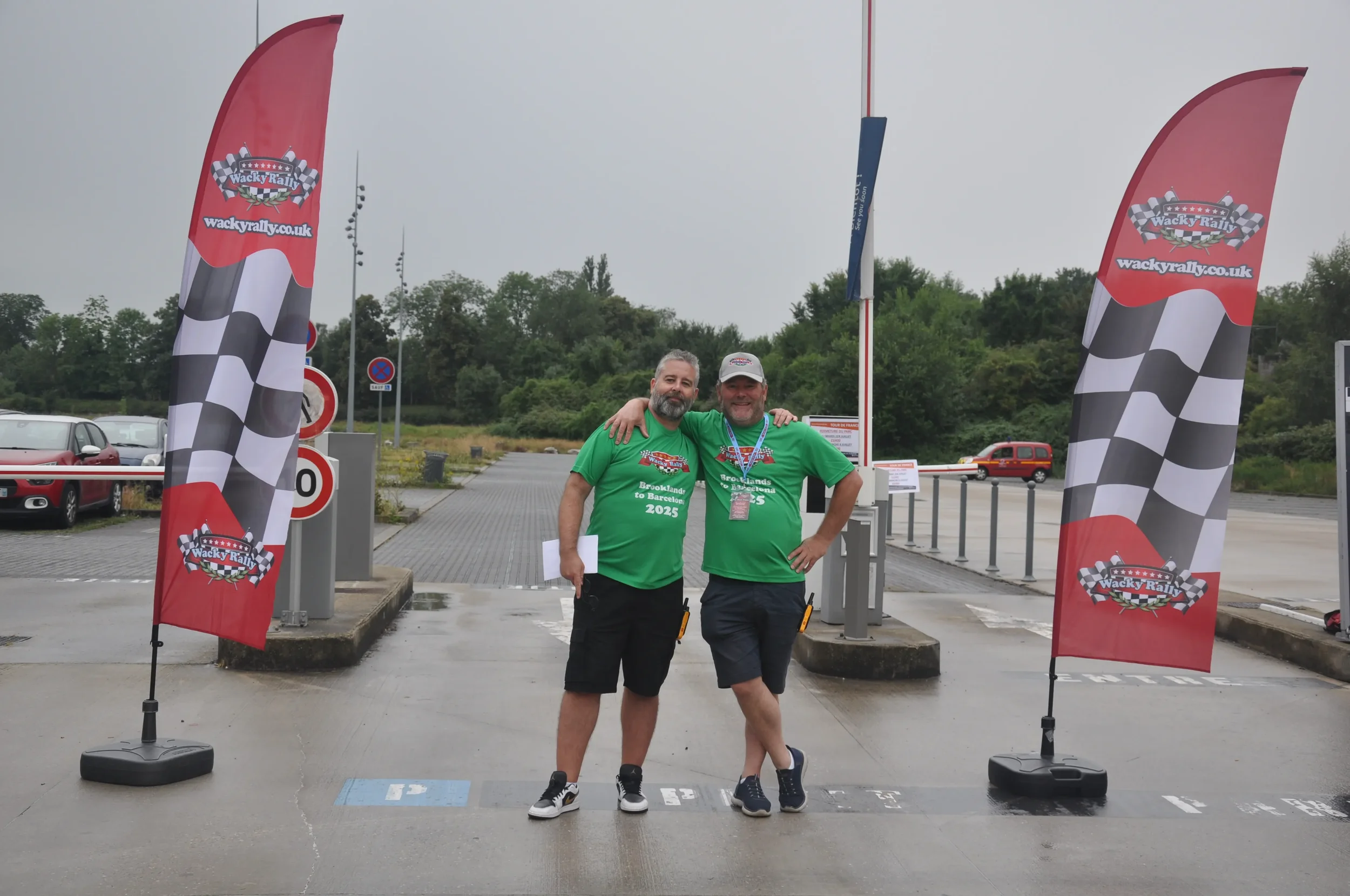 Two men standing together with arms around each other in front of a checkered flag banner, at a racing event, wearing green shirts that say 'Brooklands to Barcelona 2025'.