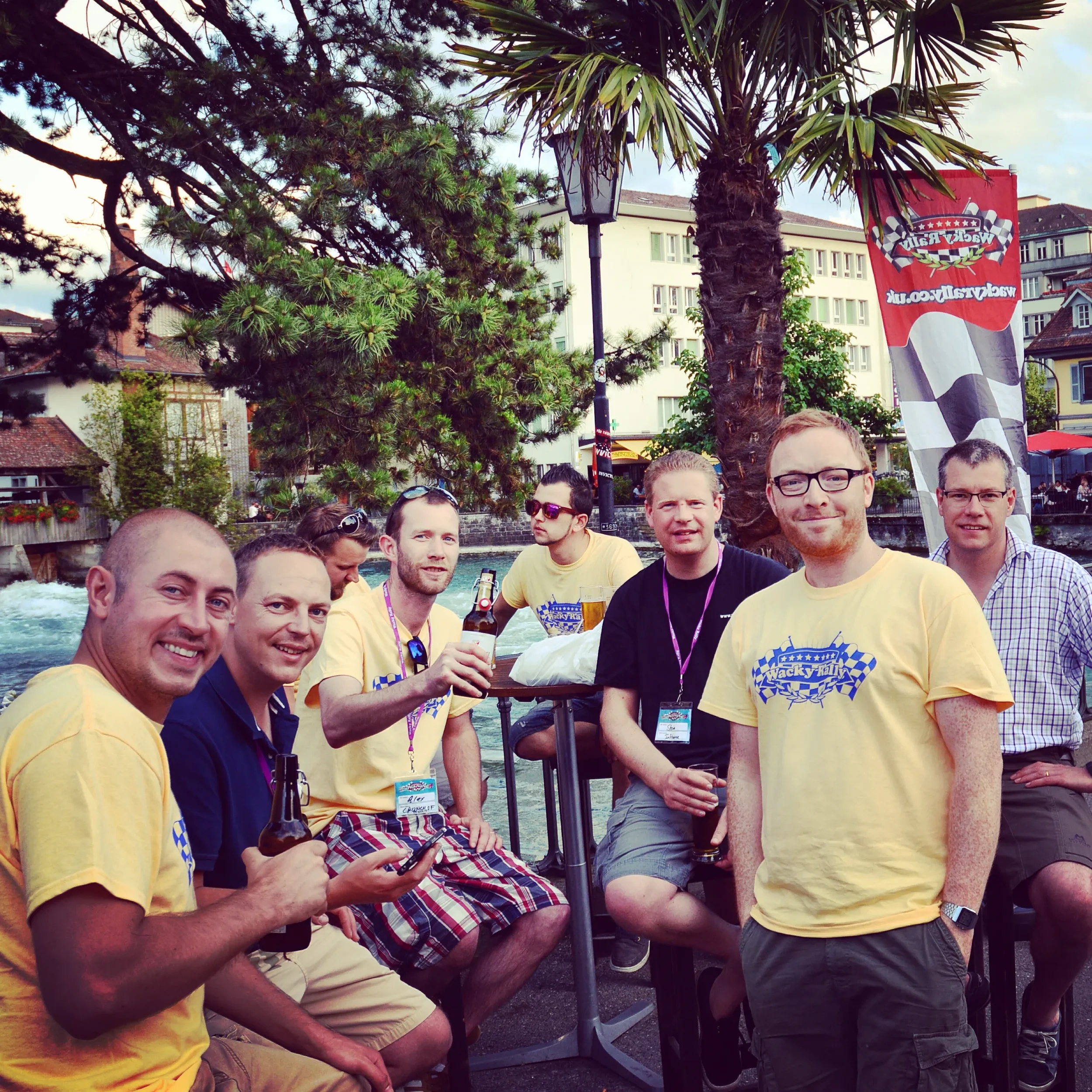 Group of eight men gathered outdoors near a river, with trees and buildings in the background, some holding drinks and smiling at the camera, during a sunny day.