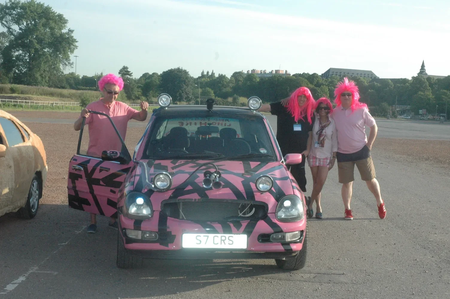 Group of four people wearing pink wigs standing outside a pink and black car, all smiling, in an outdoor parking lot on a sunny day.