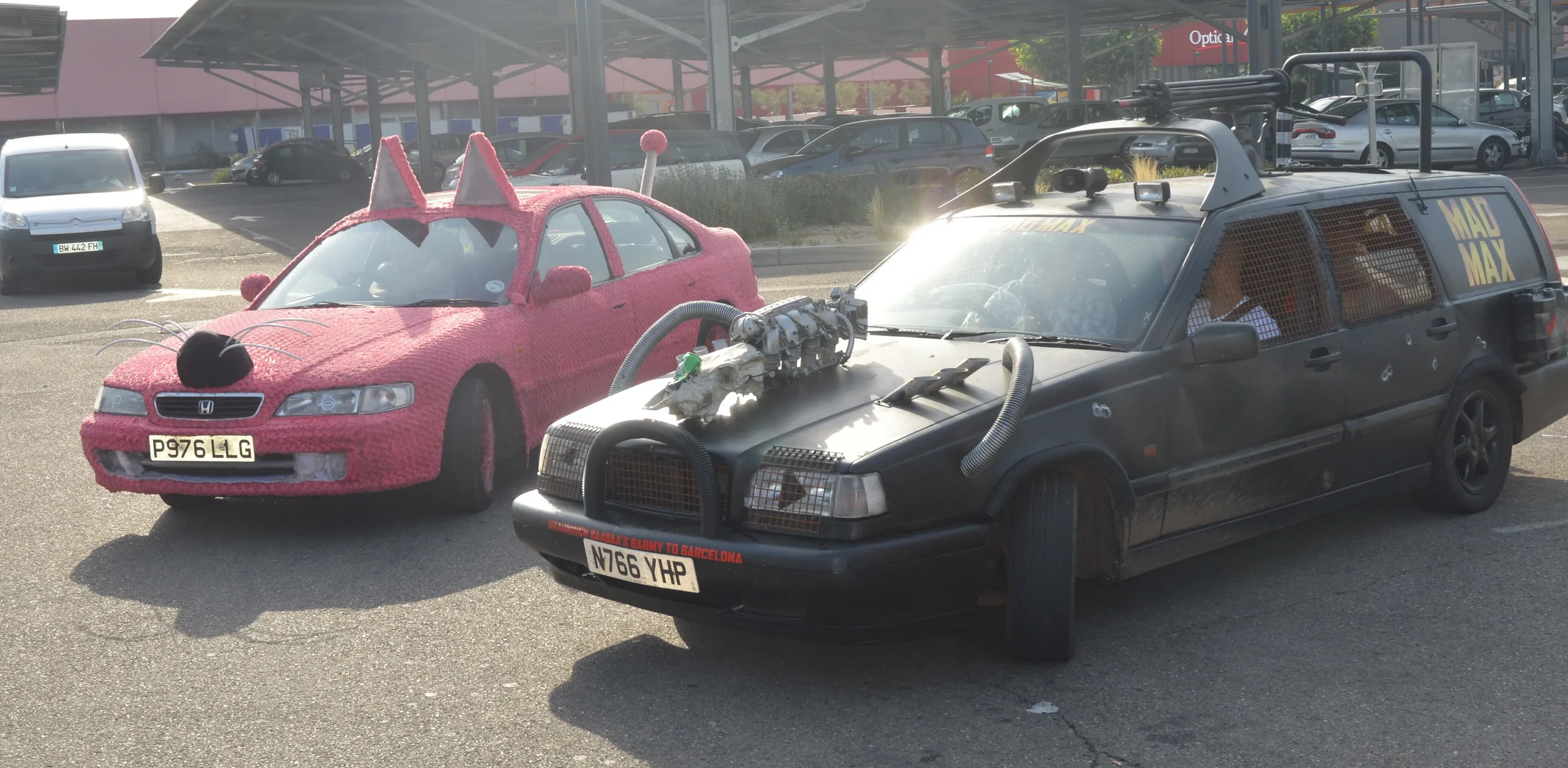 Two cars parked side by side: one decorated in pink with cat-like features, and the other designed to look like a fictional vehicle with prop weapons and a matte black finish.