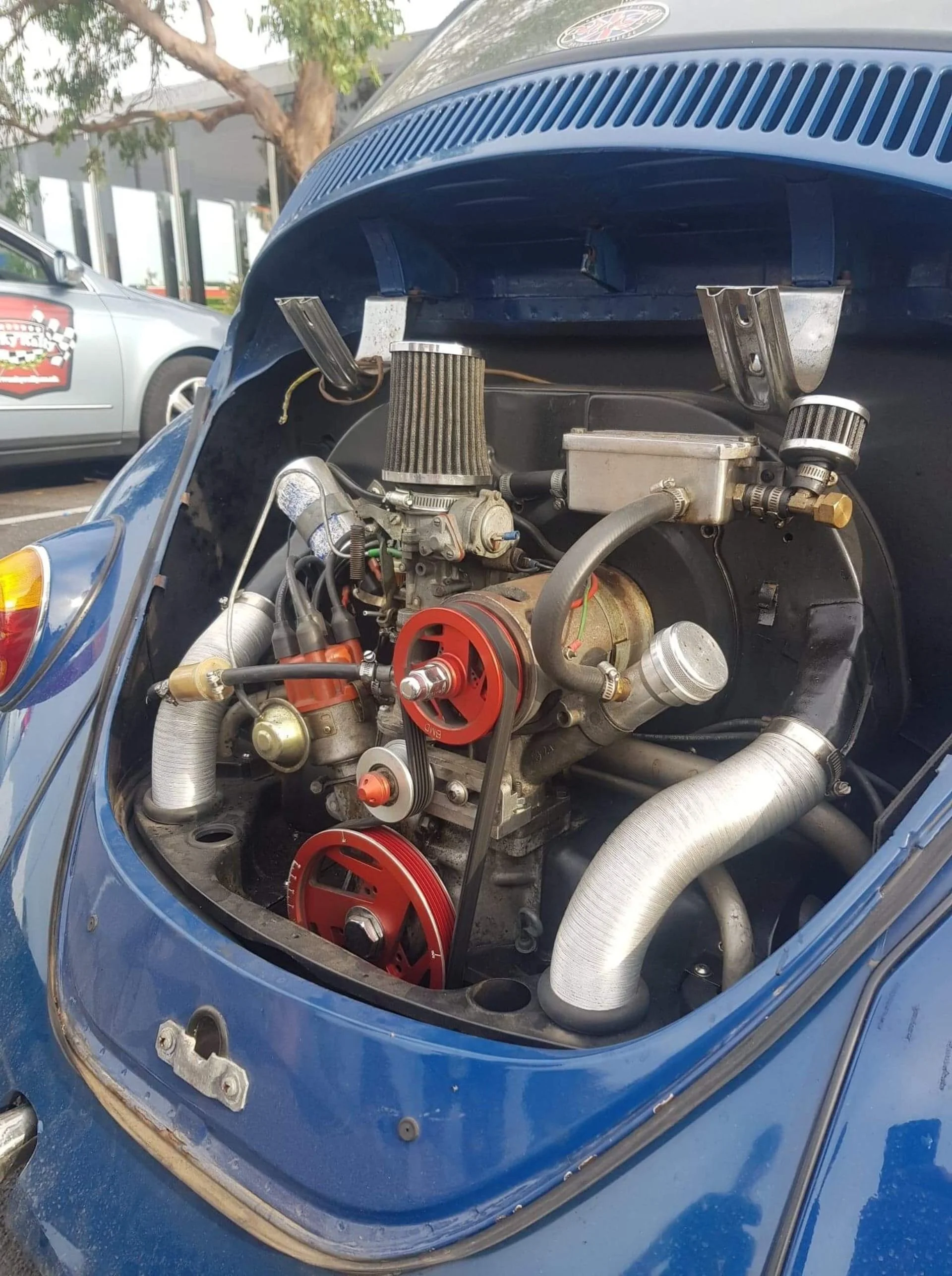 Close-up of a vintage blue Porsche with the engine hood open, exposing the engine with visible red pulleys, belts, and various mechanical parts.