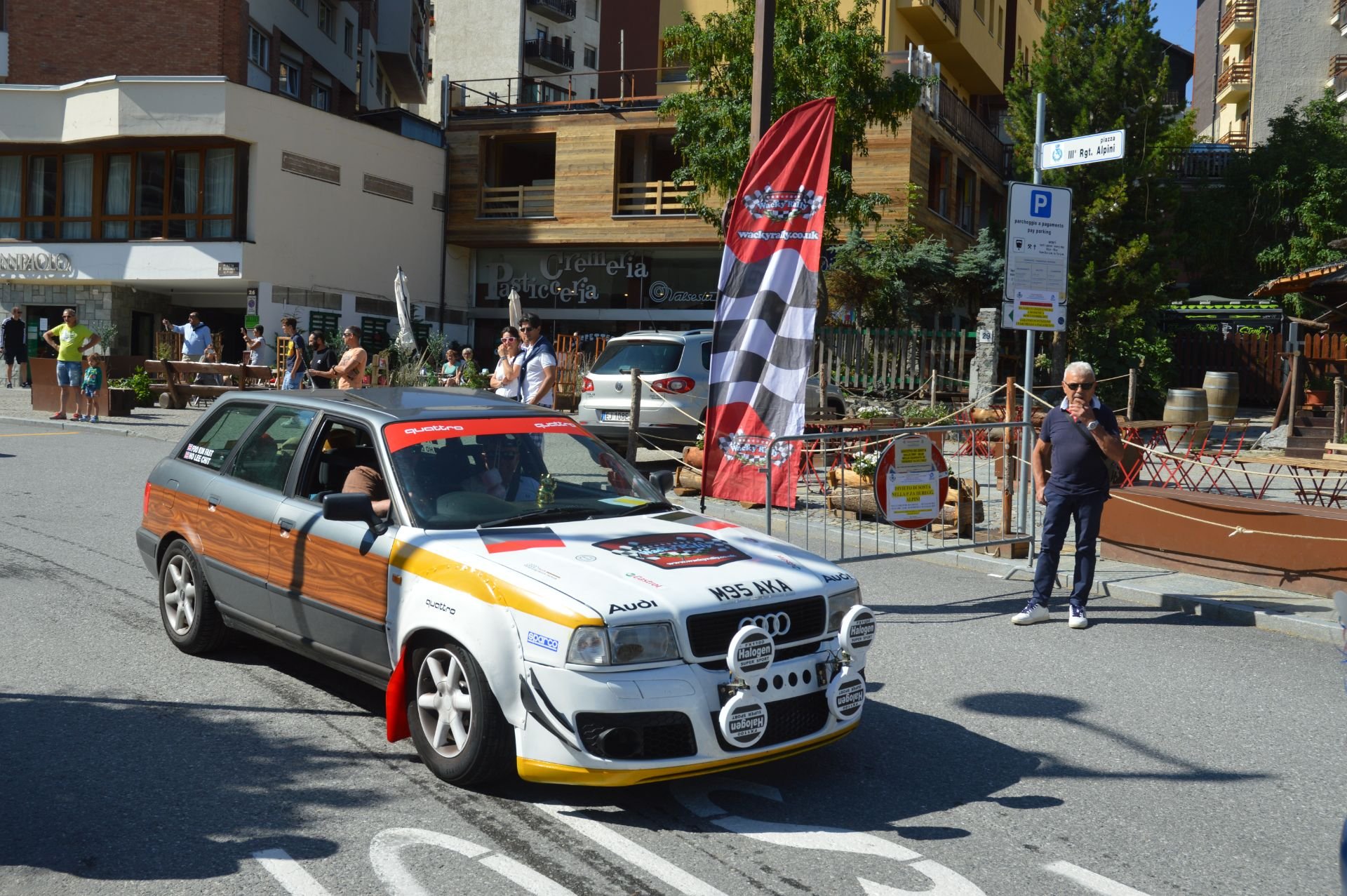 A vintage Audi rally car with sponsor decals and halogen lights is driving on a city street during a car event, with people watching from the sidewalk.