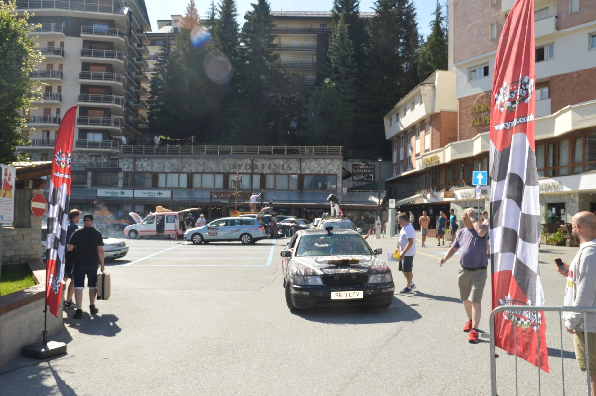 A parking lot in front of a building with the sign 'Sportiva' with several cars, people walking, and two red flags with a checkered pattern and logos.
