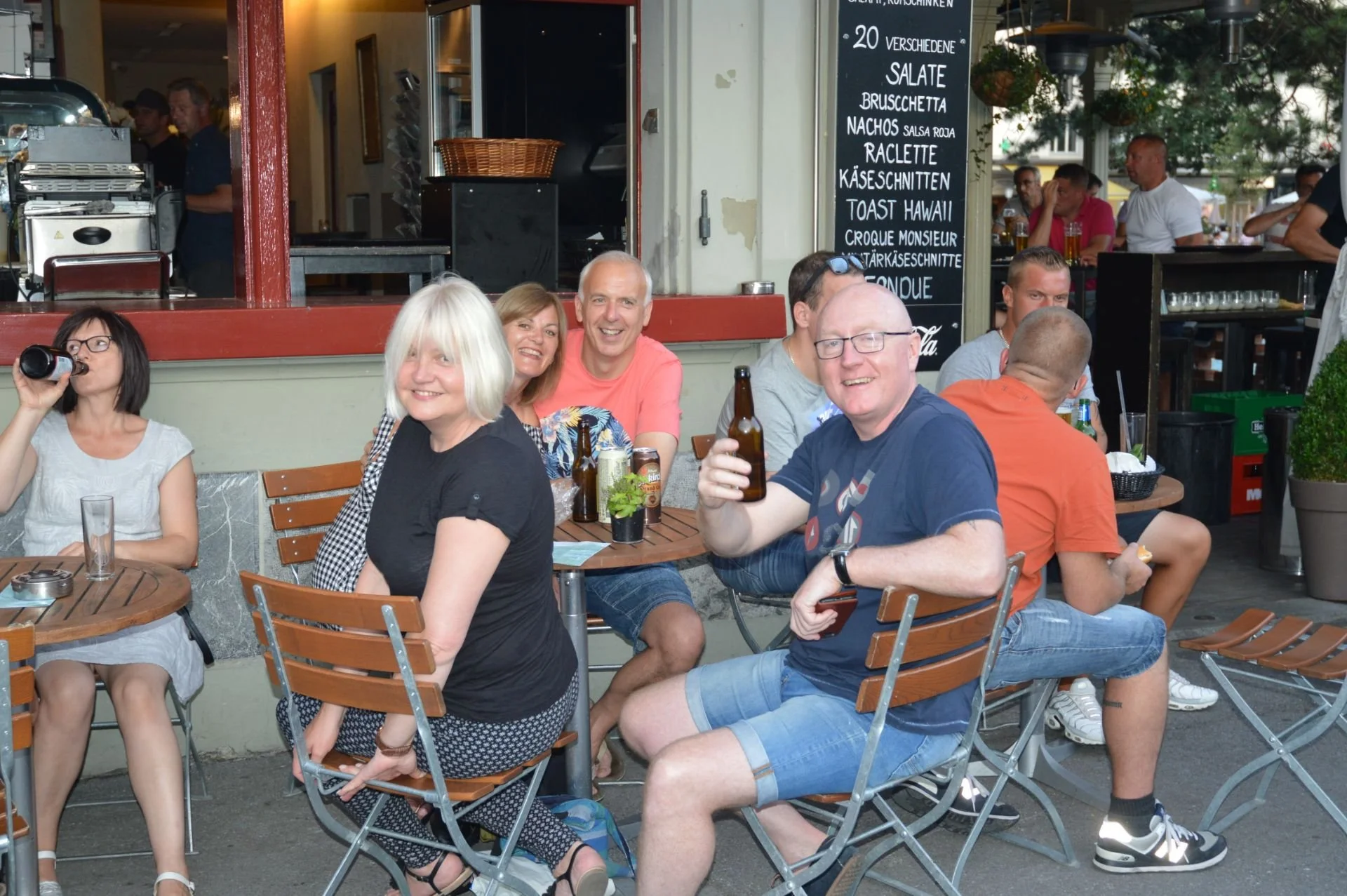 Group of people sitting at a table outside a restaurant, smiling and holding drinks, with a menu board in the background listing various food items.