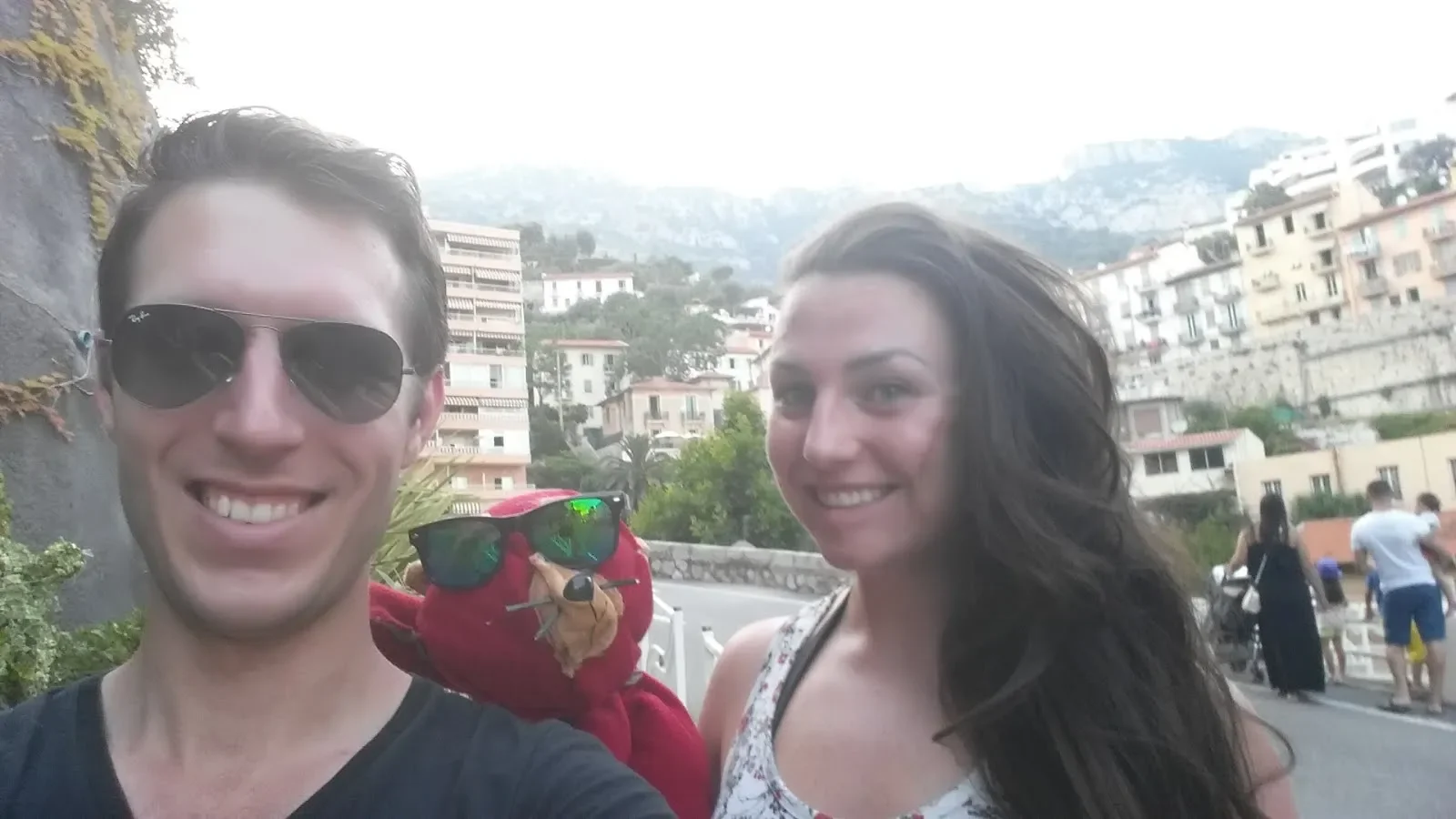 A smiling young man and woman taking a selfie outdoors in a hilly, urban area. The man is wearing sunglasses and a black shirt, and the woman has long dark hair and a sleeveless top. In the background, there are people, buildings, and hills.