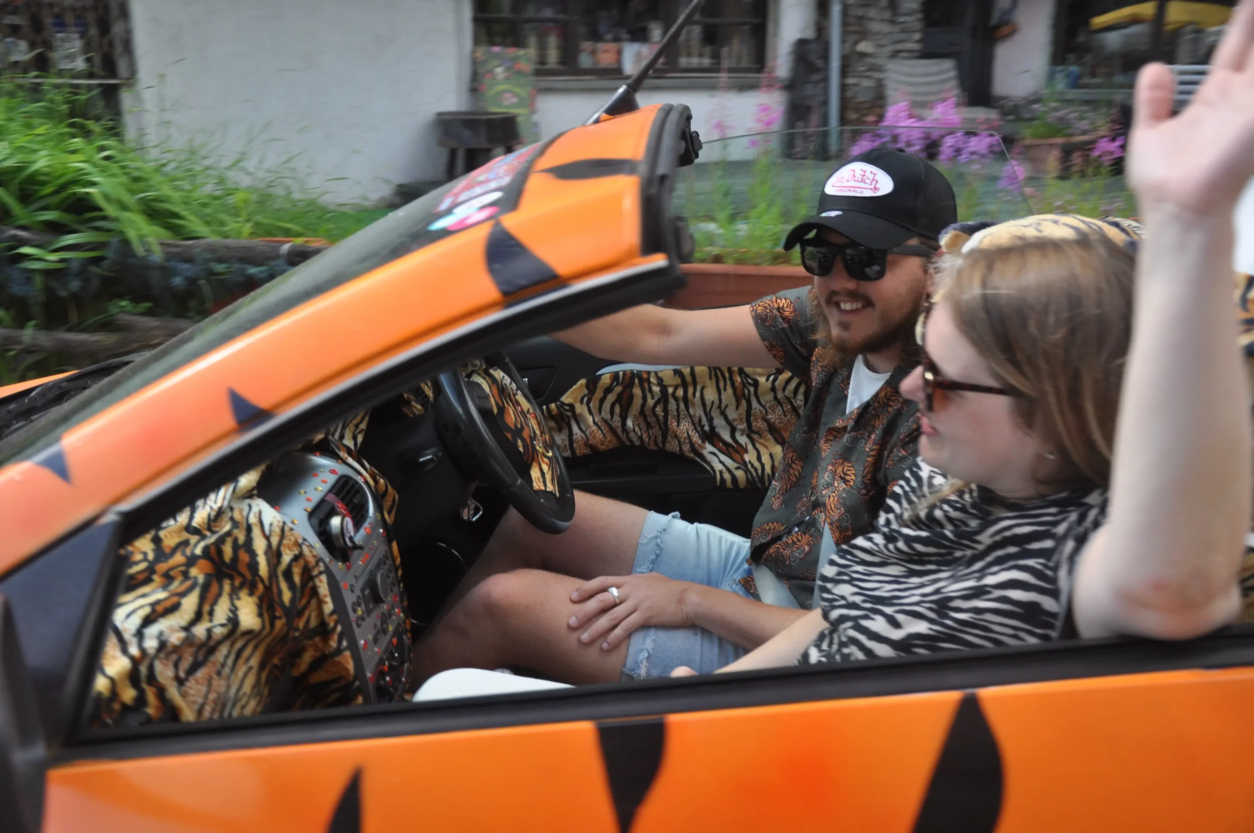 A man and a woman sitting in a small orange car with a tiger stripe pattern. The man is wearing sunglasses, a cap, and a tropical shirt, smiling and waving. The woman has glasses, a zebra striped shirt, and is looking out of the window. The backgroun