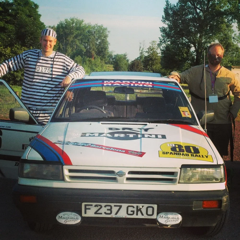 Two men standing next to a white rally car with racing decals, one wearing a striped outfit with a number badge, and the other wearing sunglasses and a mask, posing outdoors with trees in the background.