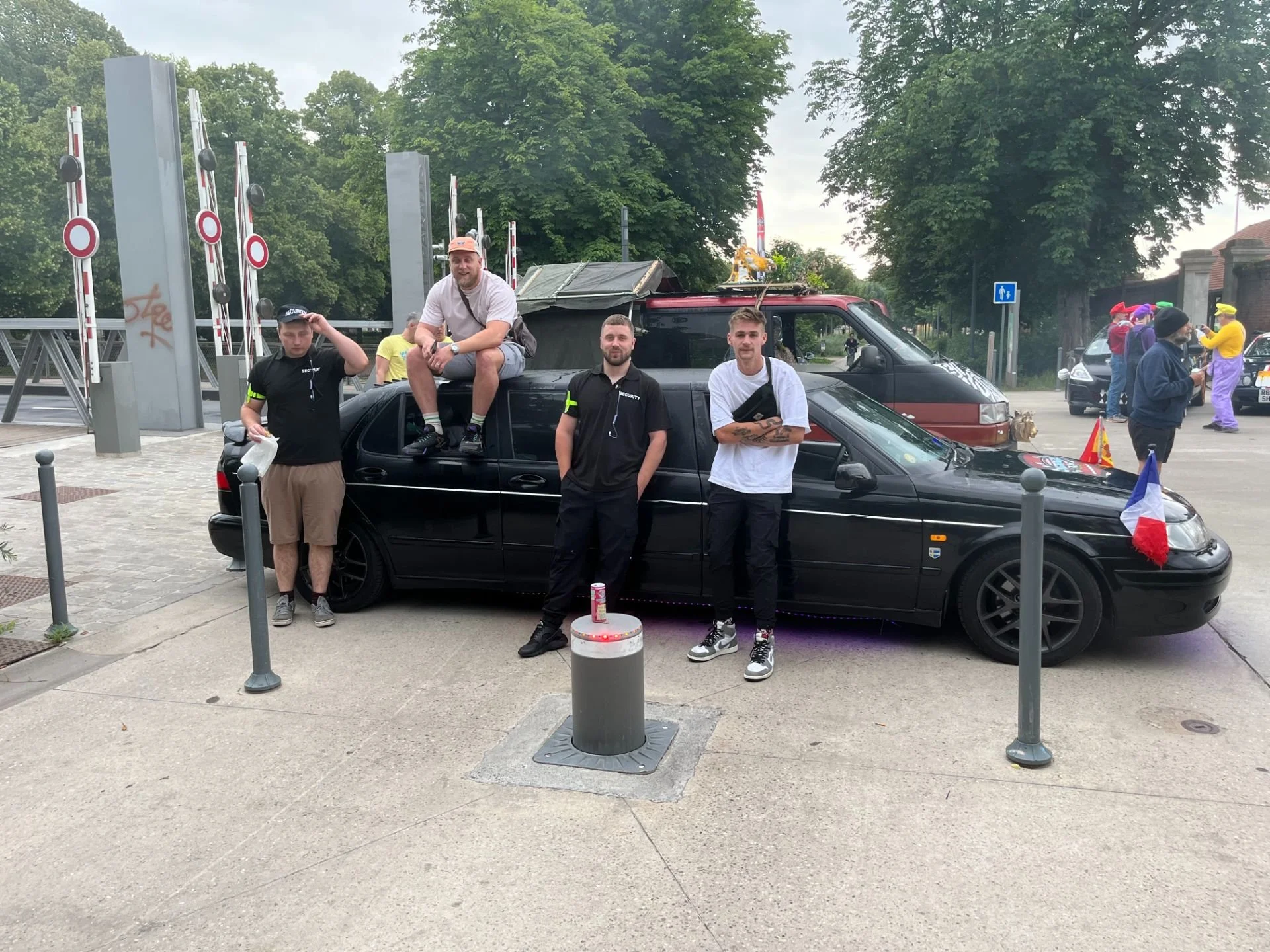 Four people posing next to a black car decorated with French flags, with a group of clowns in the background on a city street.