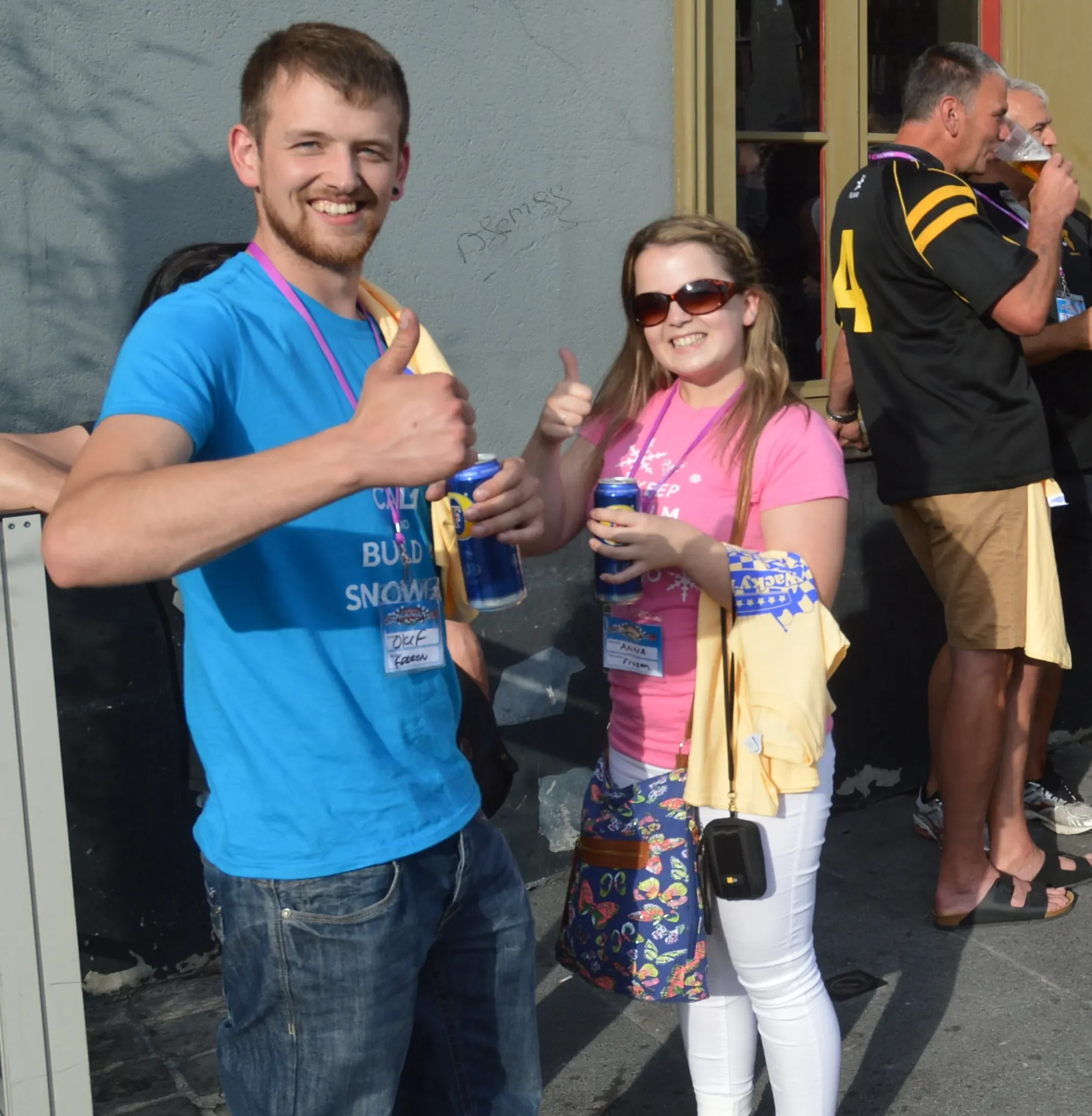 Two young people smiling and giving thumbs up while holding cans of drinks, at an outdoor event with other people in the background.