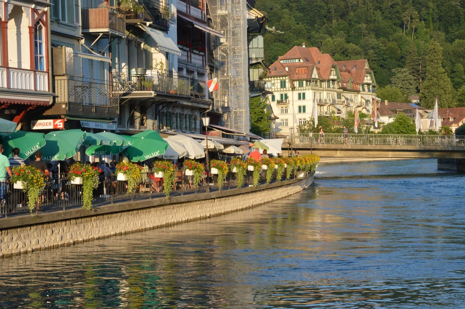 A scenic view of a riverside walkway lined with buildings, outdoor cafes, green umbrellas, and colorful flowers, with a mountain in the background.