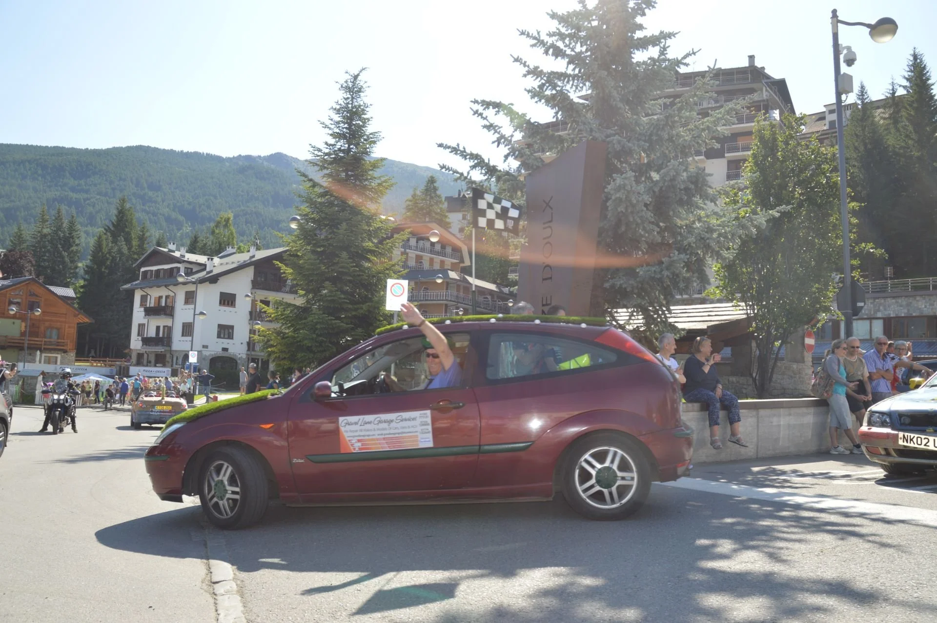 A man waving from a red car parked on a street in a mountain town, with other people sitting on a wall and buildings, trees, and mountains in the background.