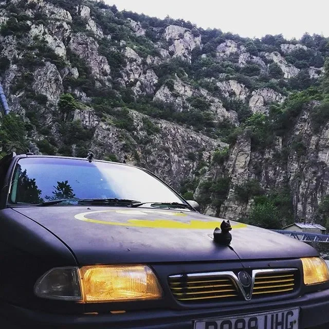 A black car with a yellow Batman logo on its hood is parked outdoors against a rocky mountain background. A small black cat is sitting on the hood.