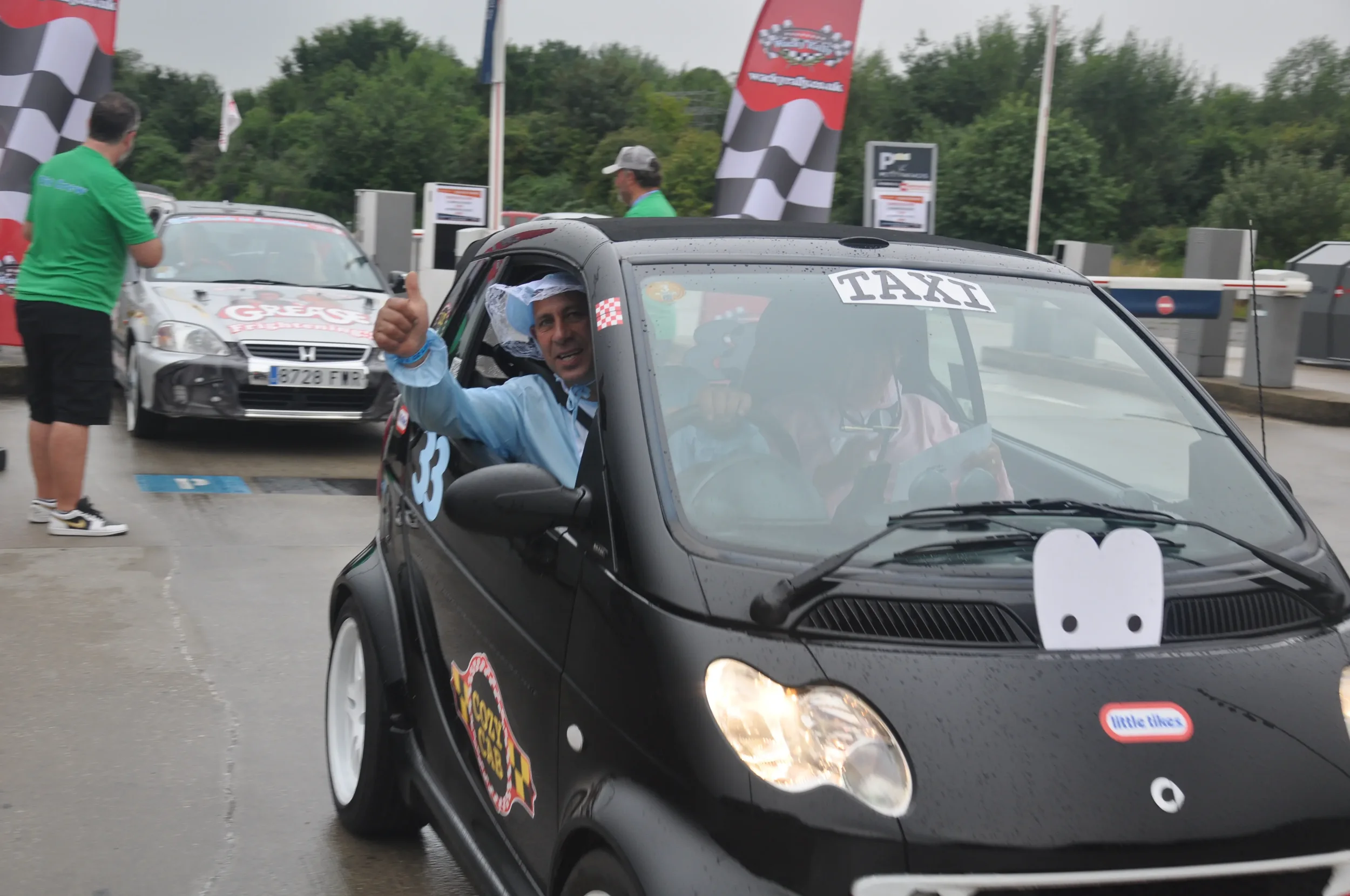 Man in a blue jacket giving a thumbs-up while sitting in a black Smart car with race stickers, at a rally or racing event with other cars and people in the background.
