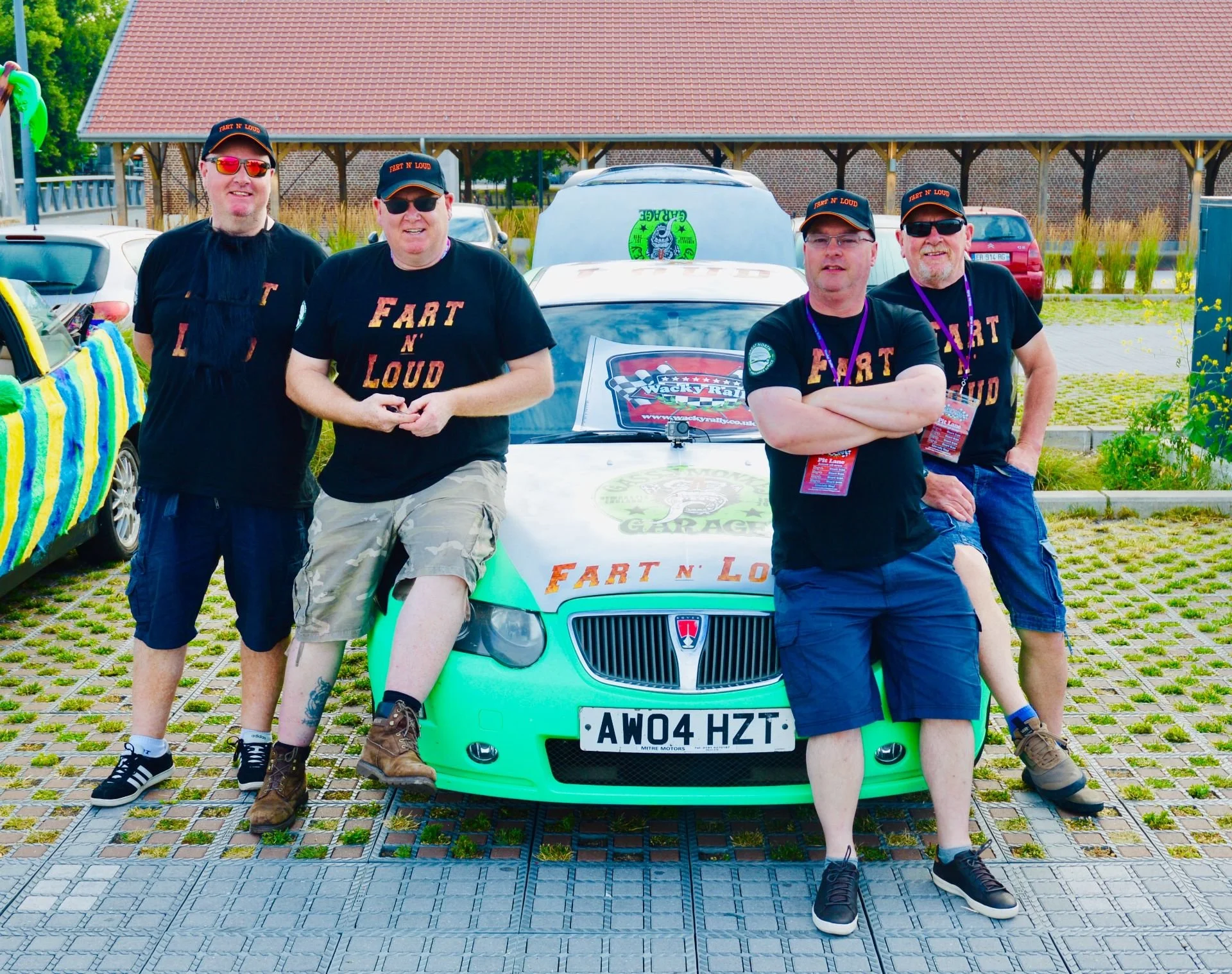 Four men posing next to a car at an outdoor event, all wearing black t-shirts with yellow and orange text that reads "Fart N Loud," and caps. The car is decorated with stickers and the license plate reads "AW04 HZT."