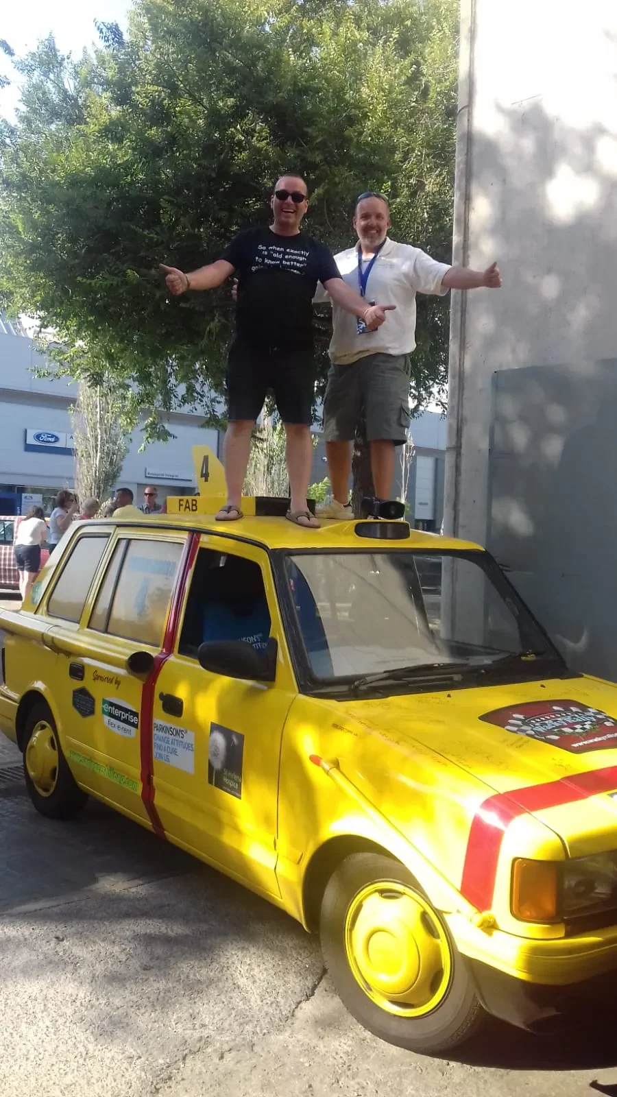 Two men standing on top of a yellow race car with sponsor stickers, smiling, with thumbs up, in an outdoor area with trees and people in the background.