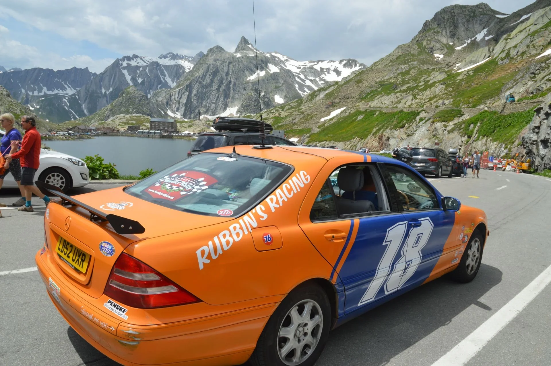 A colorful race car with orange and blue paint, numbered 18, parked near a mountain lake with snow-capped peaks in the background. There are people walking and parked cars along the road.