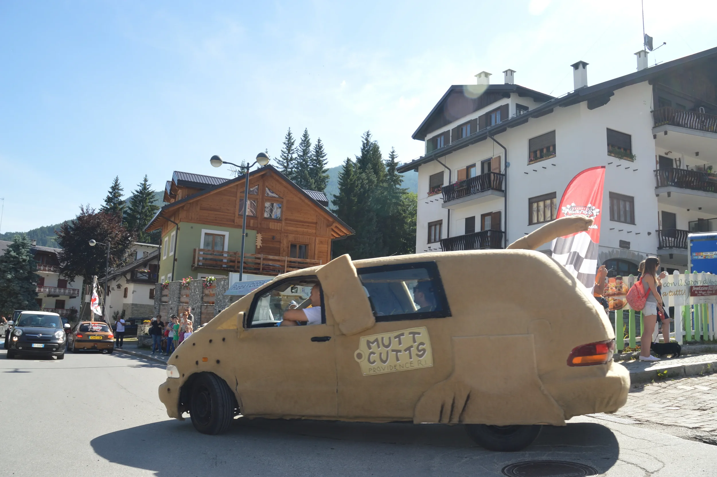 A person driving a car covered in cardboard resembling the 'Mod' style of The Flintstones, with a sign that reads 'Mutt Cuts' on the door, in a street with colorful buildings and people in the background.