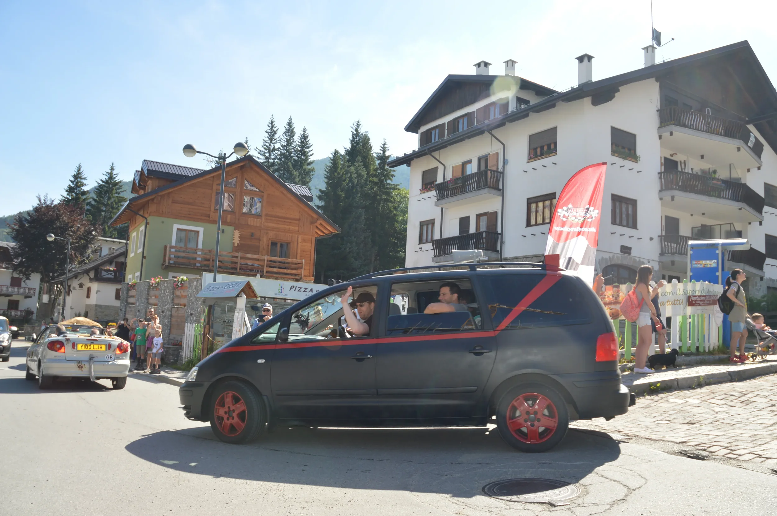 A black car with red rims parked on a city street, with a man waving to someone inside, surrounded by people, buildings, and trees, in a mountain town.