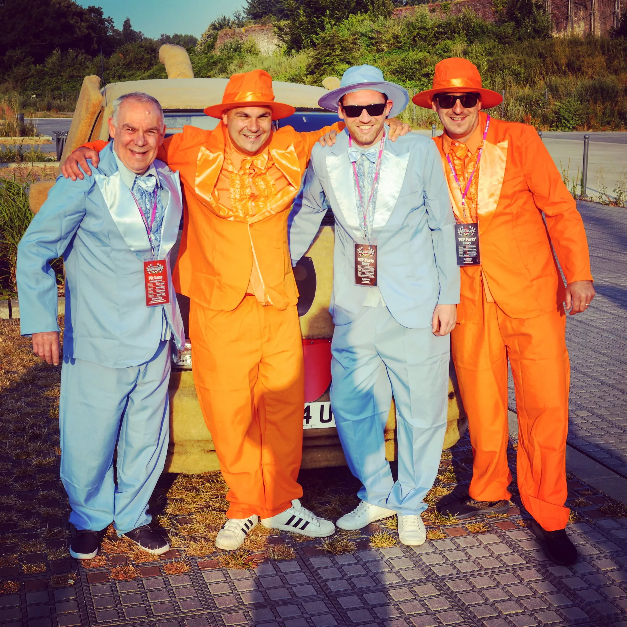 Four men dressed in colorful suits and hats, standing outdoors in front of a vehicle, smiling, with one in a blue suit, two in orange suits, and one in a light blue suit, during daytime.
