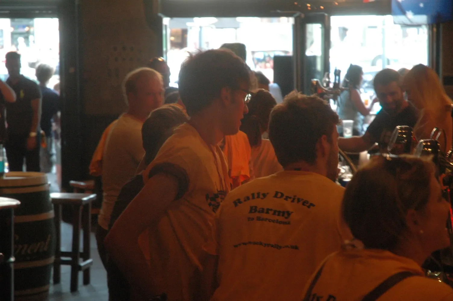 A group of people in yellow shirts gathered at a bar or pub, some are sitting and some are standing near the counter, with a bar tap system visible in the foreground. The background shows more patrons and glass doors or windows letting in daylight.