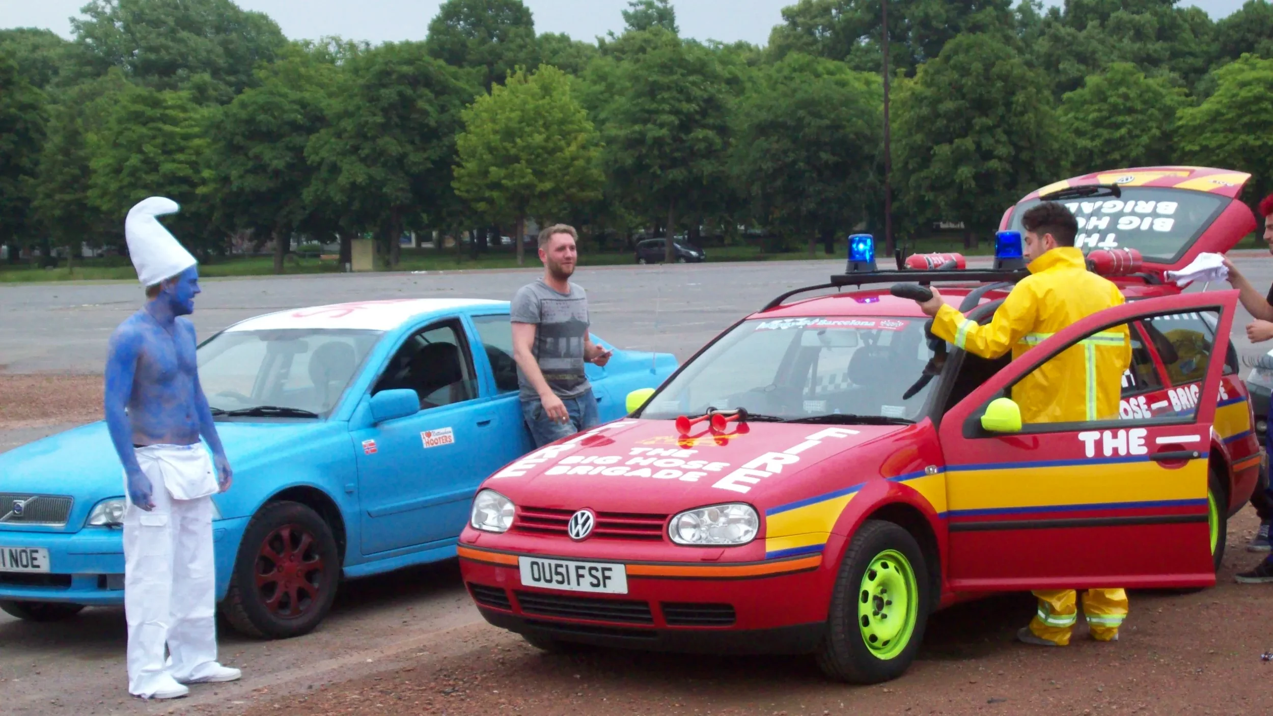 A person dressed as a Smurf with blue body paint, white pants, and a tall white hat, standing next to a blue car. Another person is holding a phone, and a firefighter in bright yellow protective gear is inside or beside a red emergency vehicle with f