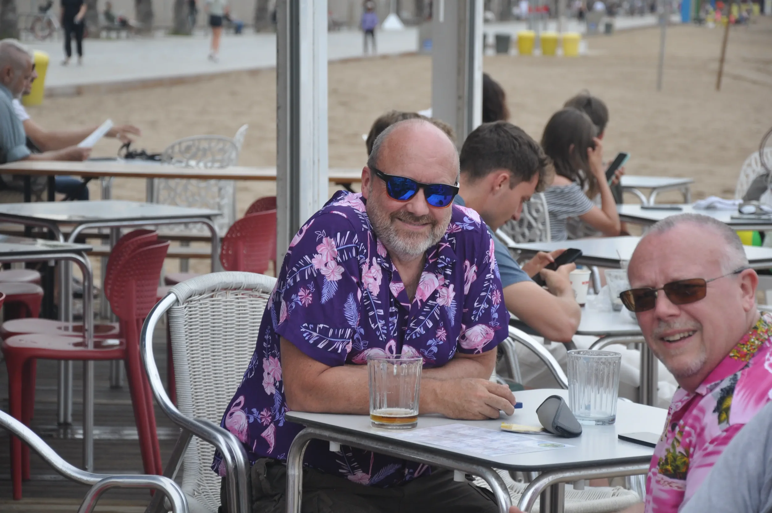 Two men wearing colorful Hawaiian shirts and sunglasses sit at a table outside at a beachside cafe, smiling at the camera. Other people are seen in the background sitting and using their phones.