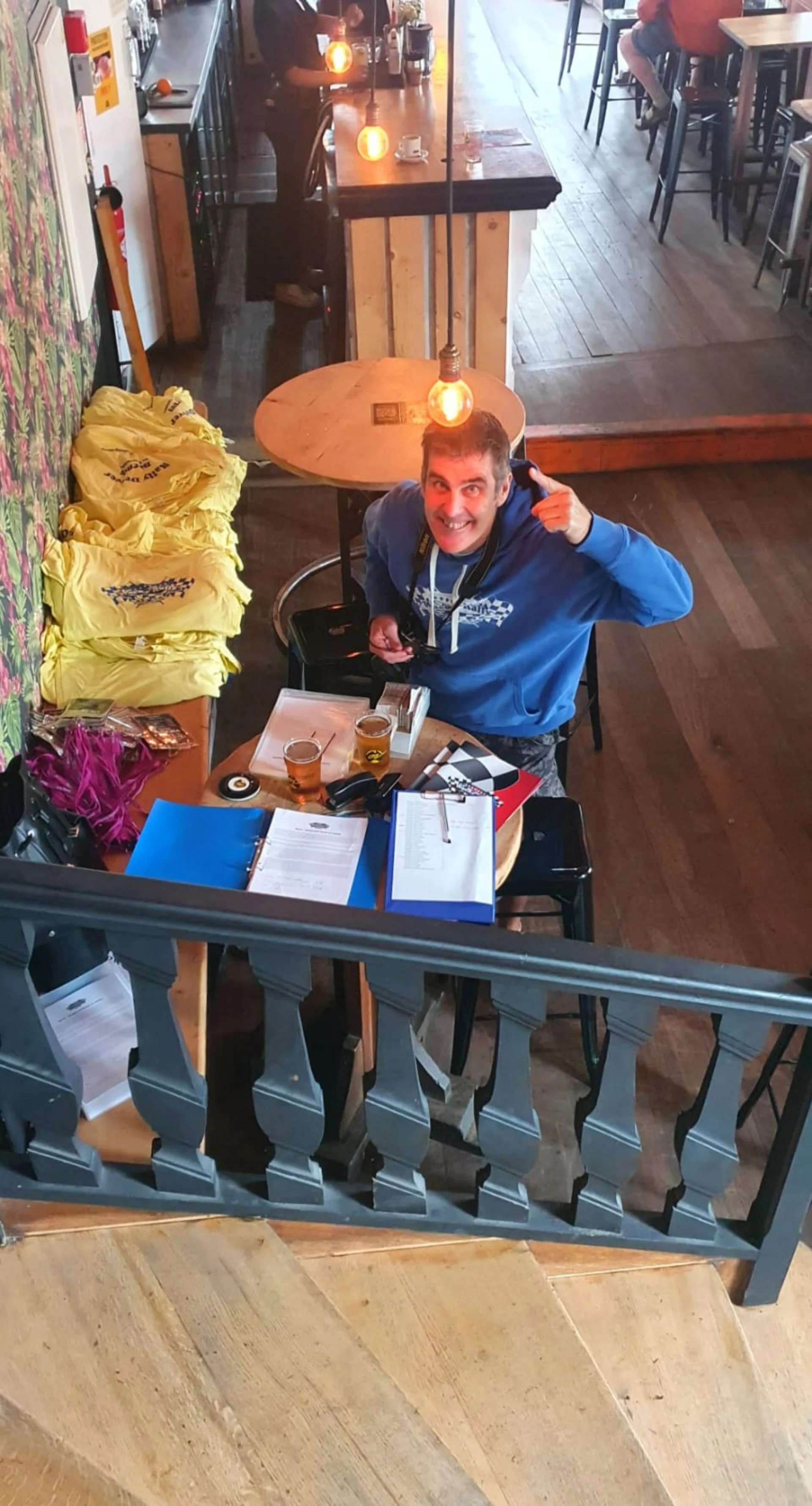 A man with a camera around his neck is sitting at a table in a restaurant, smiling and pointing at his head. The table has two glasses of beer, some papers, a pen, and a black device. Behind him, yellow T-shirts are stacked on a bench, and there are 