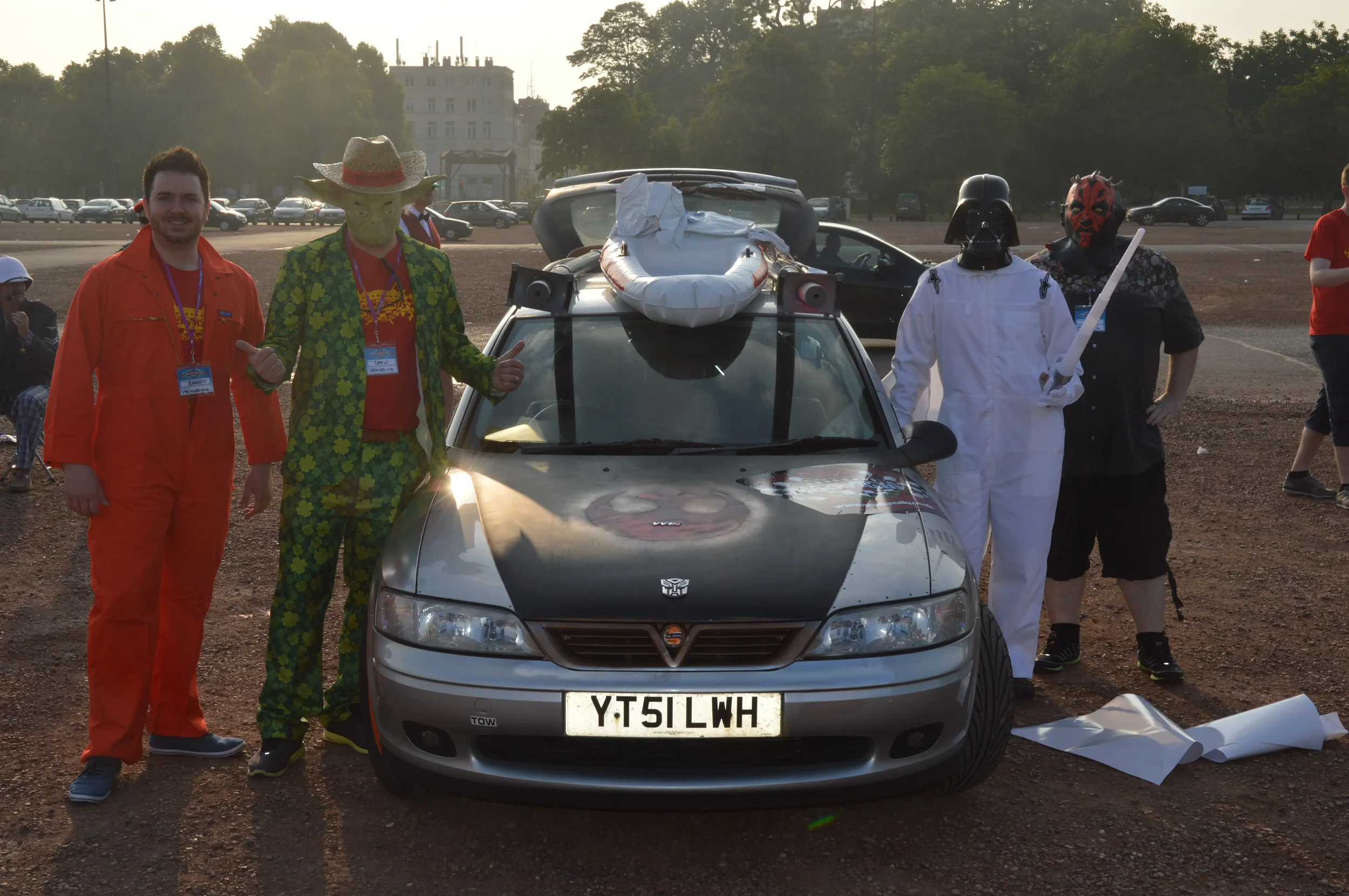 Group of five men in costumes standing next to a modified silver car with a Batman logo on the hood and a toy boat on the roof. Two of the men are dressed as Star Wars characters, Darth Vader and a Sith. The second man from the left is dressed in a c