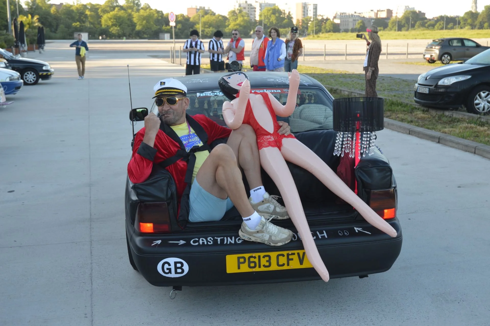 A man in a captain's hat and sunglasses sitting in the trunk of a black car with a large inflatable doll. The man is making a peace sign with one hand and holding a walkie-talkie with the other. The inflatable doll is dressed in red lingerie and is s