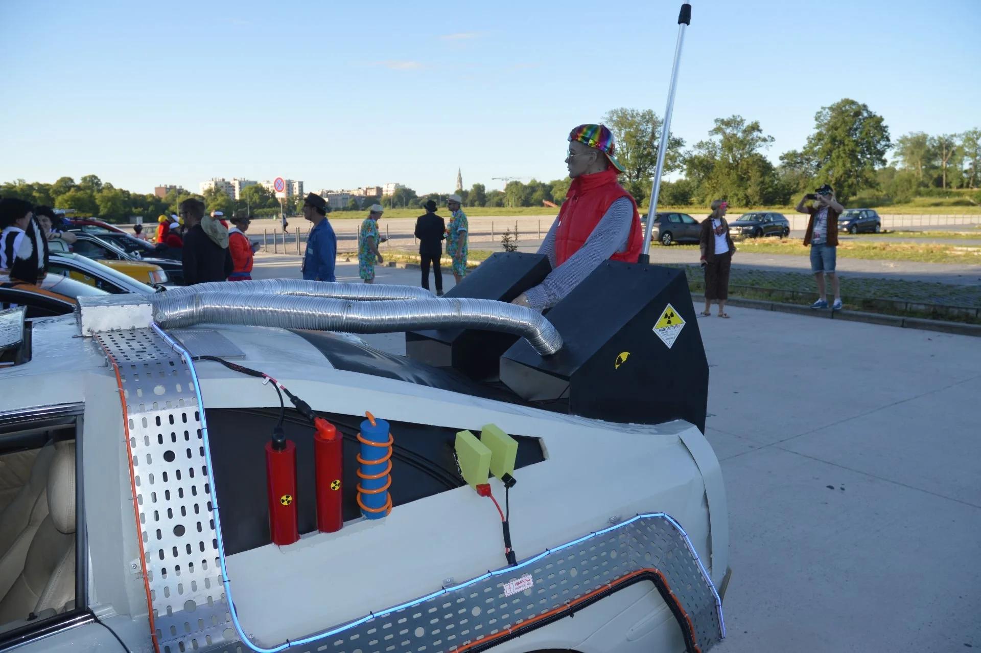 Person dressed as a 1980s-style retro-futuristic driver with metallic sunglasses and a rainbow-colored cap standing in a car with sci-fi modifications, including control panels, colored cylinders with warning symbols, and tubes, while spectators obse
