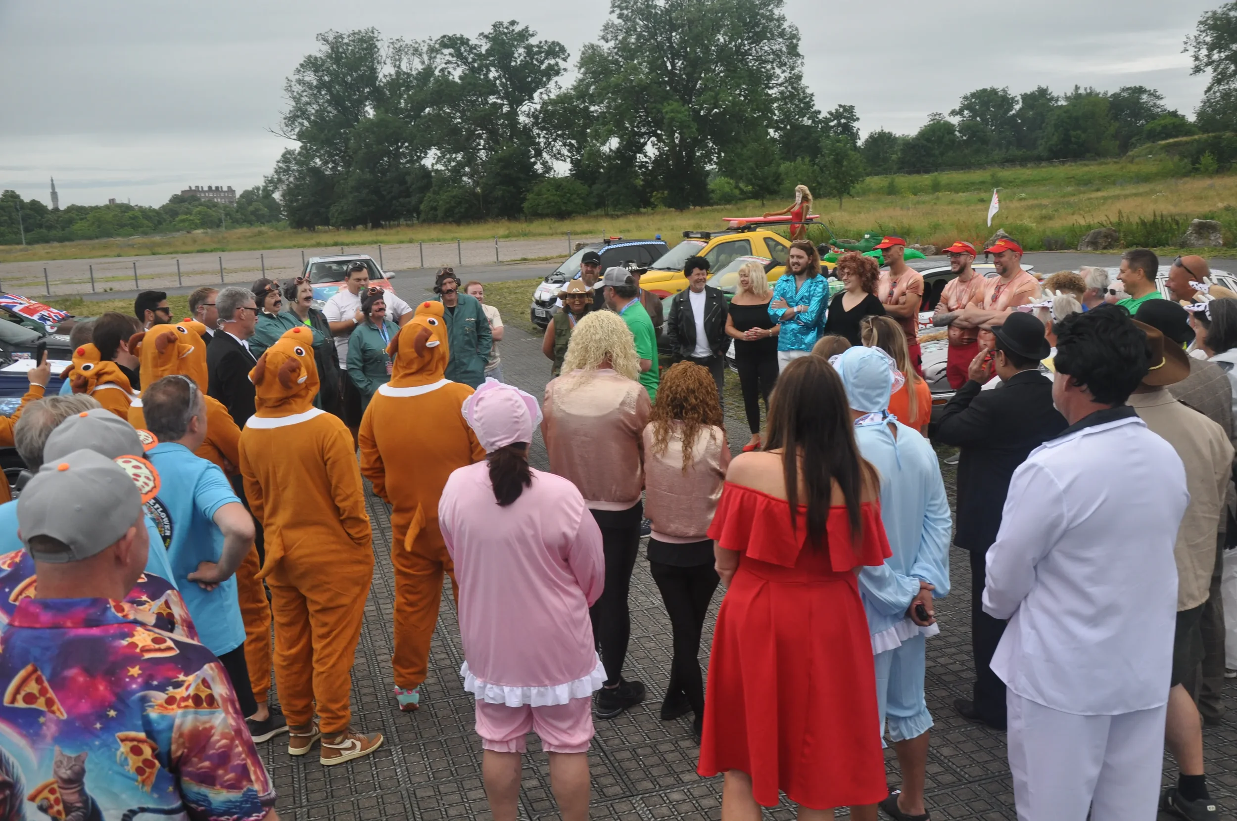A group of people gathered outdoors for a costume event, some dressed as chickens, others in various colorful outfits, with cars and greenery in the background.