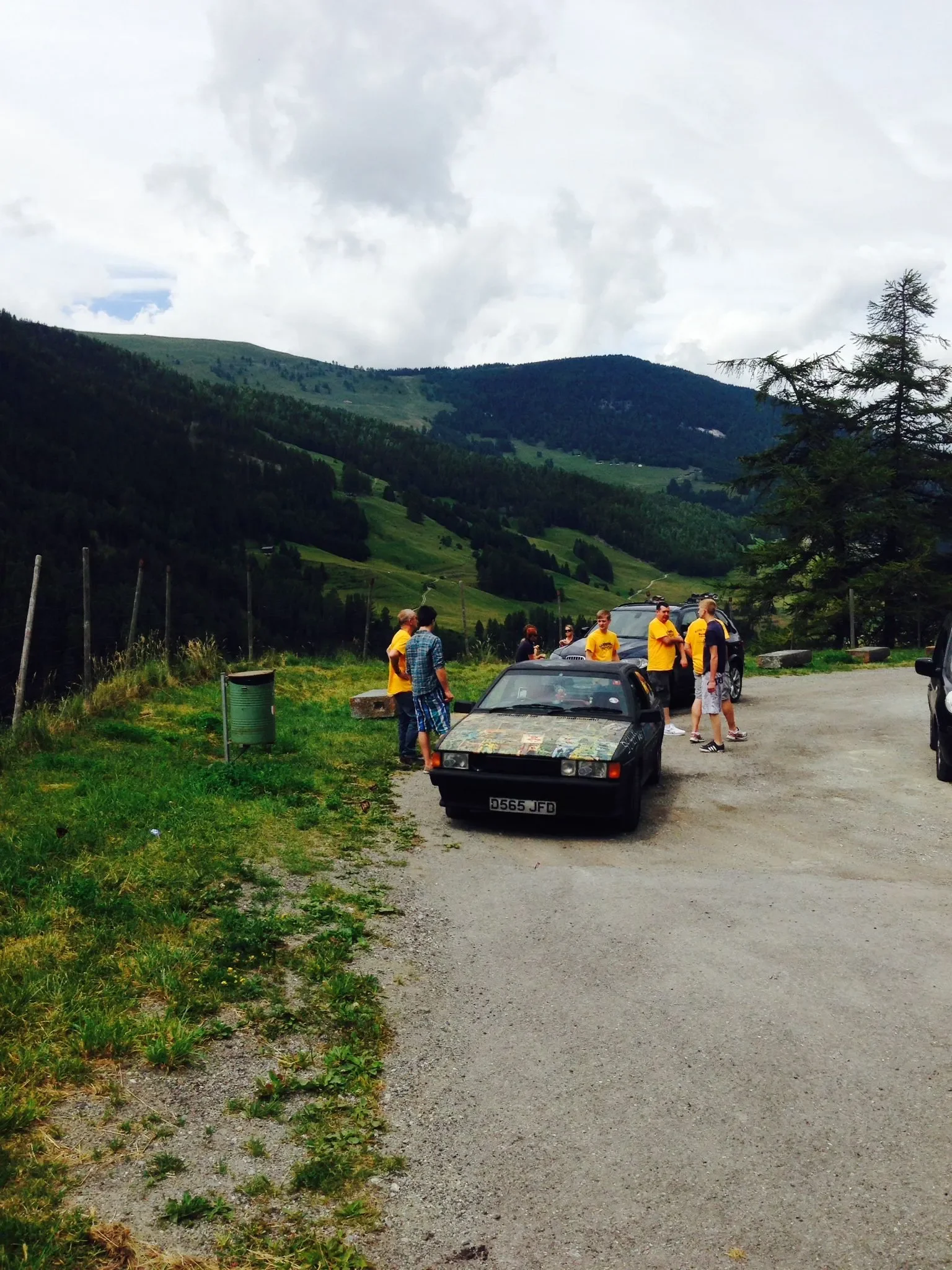 Group of people standing near a black sports car with map on the hood, in a mountainous rural area with green hills and cloudy sky.