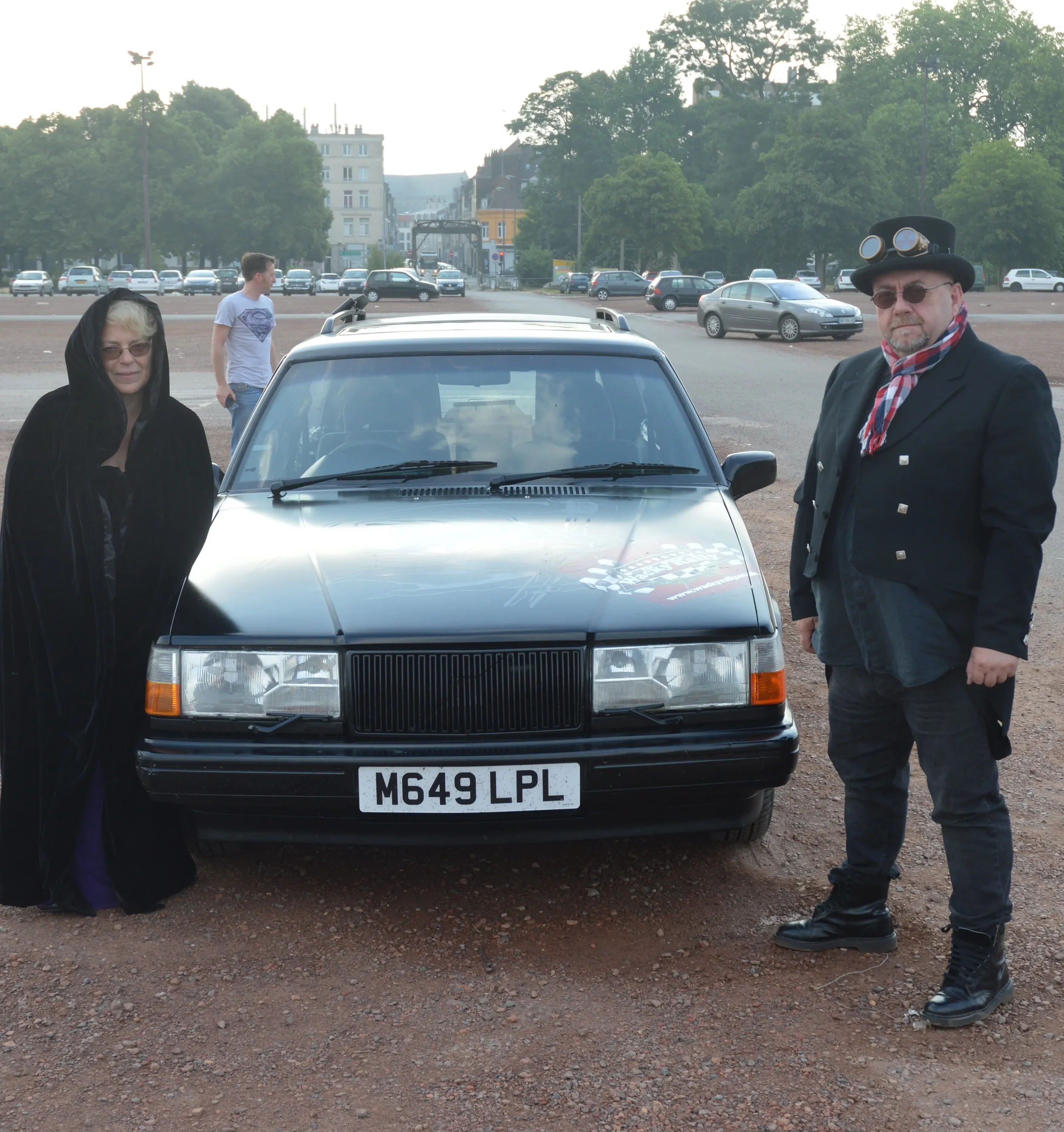 Two people standing beside a black car with a white license plate reading 'M649 LPL' in an open area, with trees, cars, and buildings in the background.