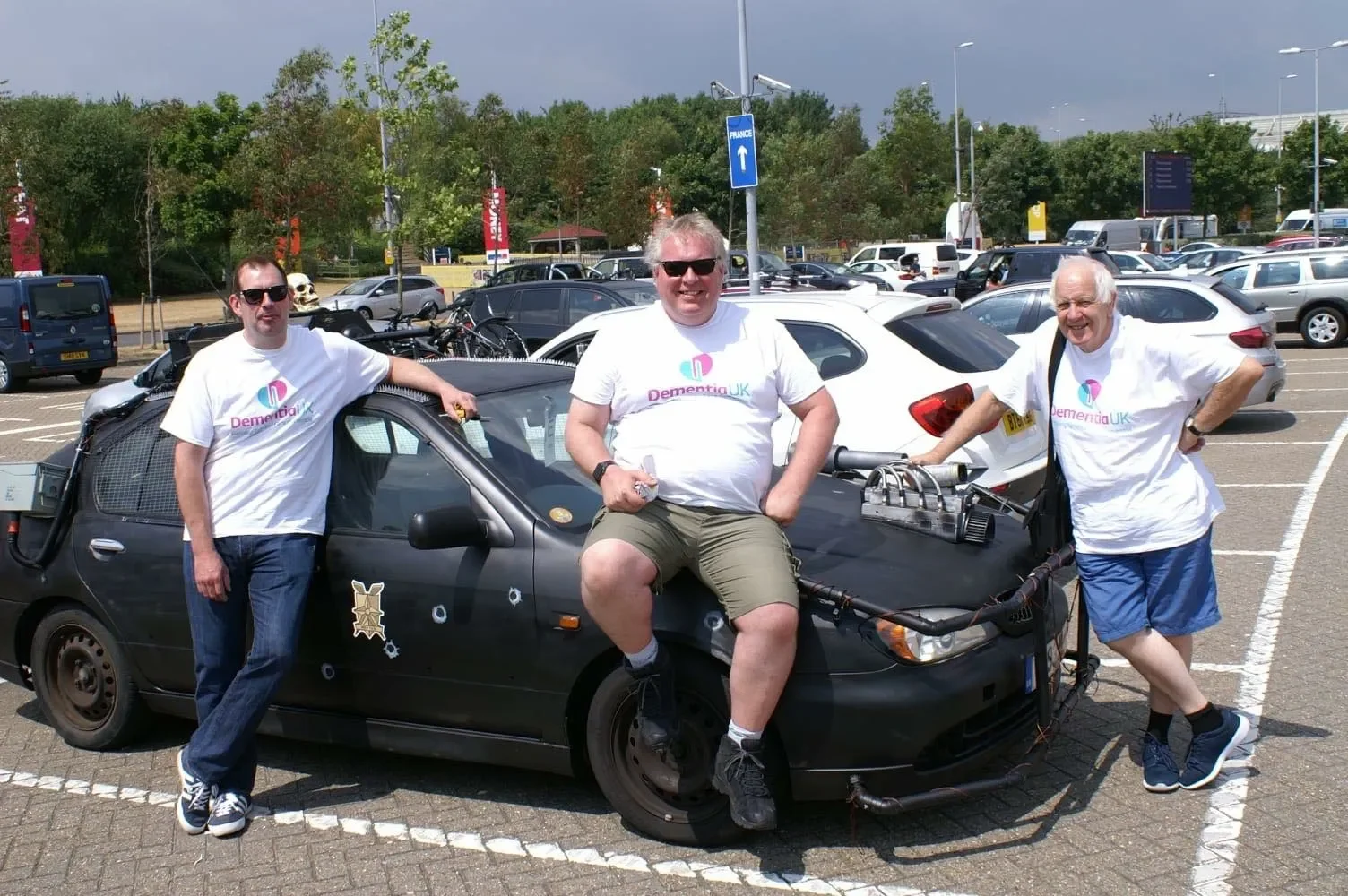 Three men standing in a parking lot next to a black car with various electronic equipment on it, smiling and wearing white T-shirts with the Dementia UK logo.