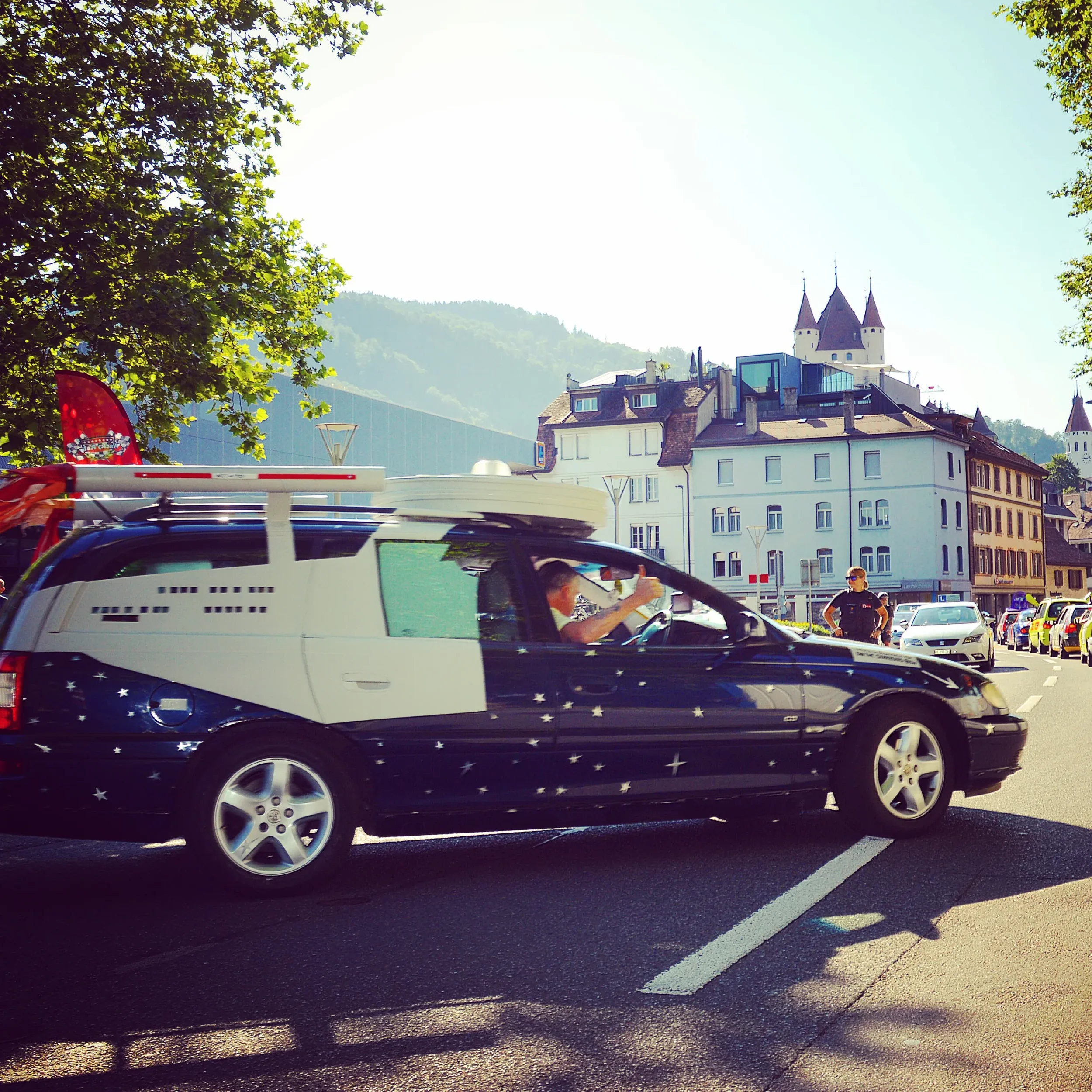 A black car with star decals and a white structure on top driving through a city street, with people and other cars visible in the background, under a bright sky with trees and historic buildings.