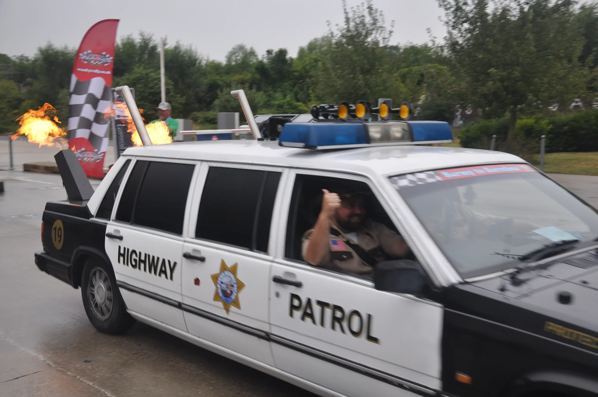 A man smiling and giving a thumbs up while sitting in a police car with a 'Highway Patrol' sign, with fire effects and checkered flags in the background, likely at a themed event or car show.
