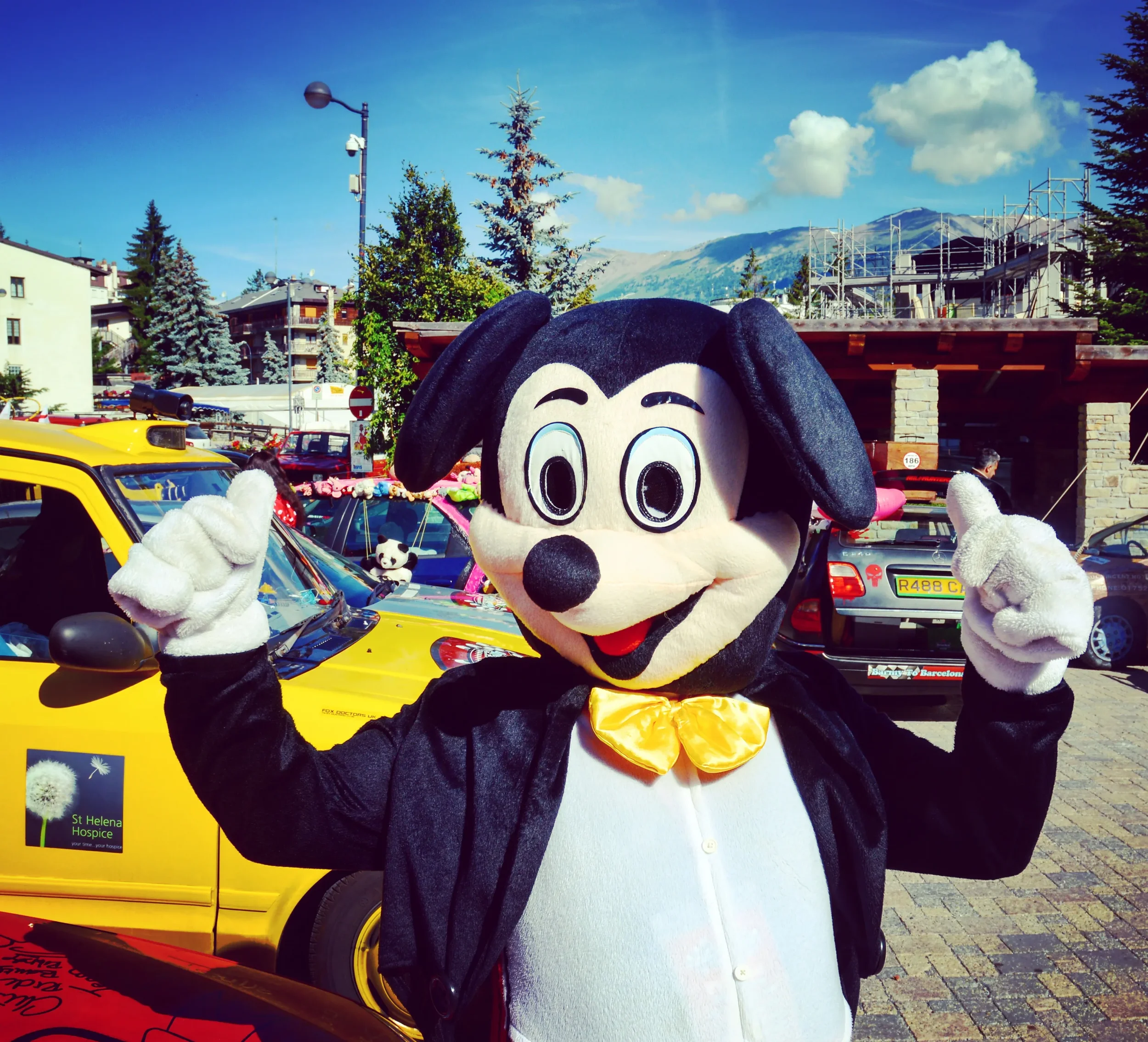 Person in a Mickey Mouse costume with a yellow bow tie, pointing upwards with both hands, standing in a parking lot with colorful cars and mountains in the background under a partly cloudy sky.