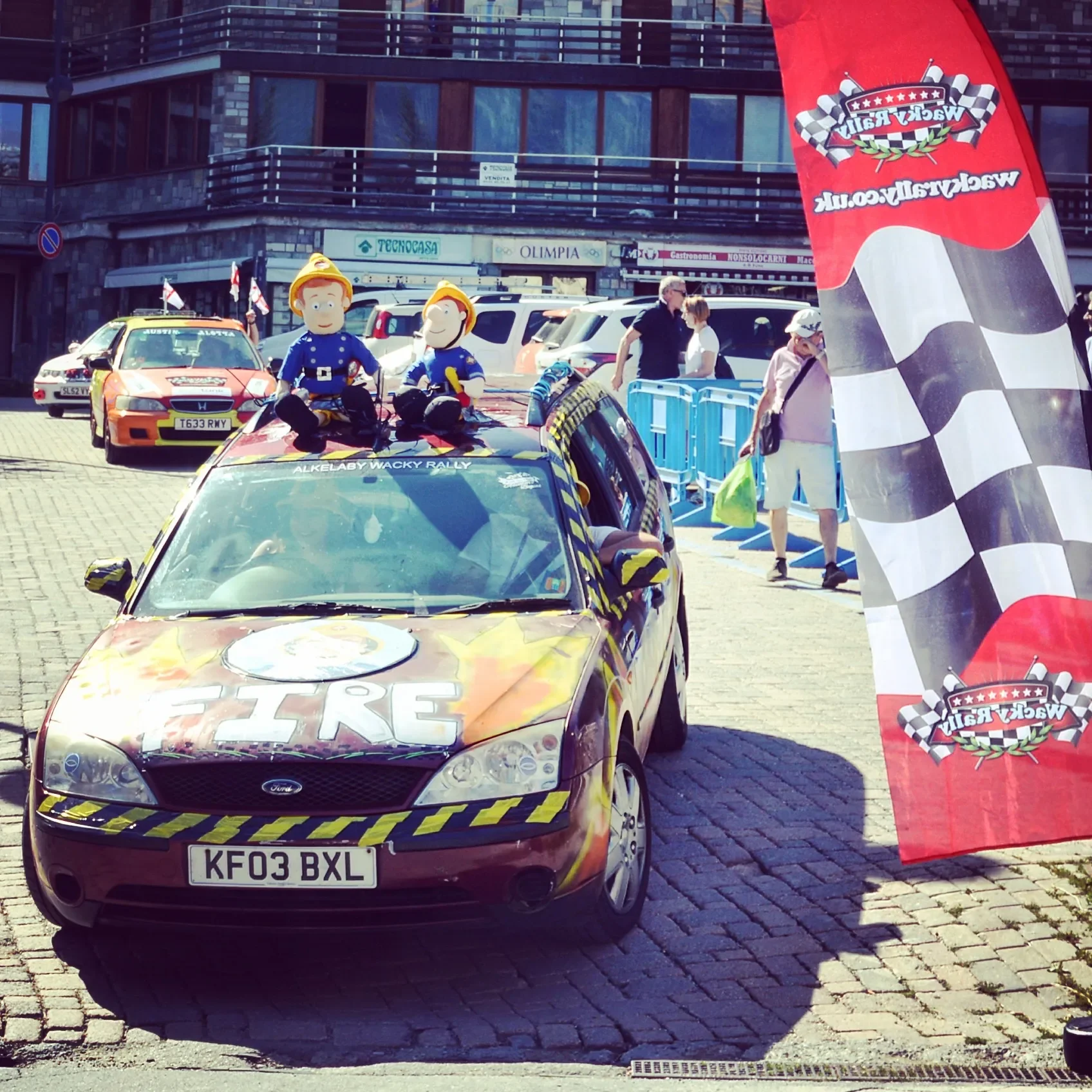 A rally car with cartoon character puppets on top, parked on a cobblestone street during a race event, with people walking nearby and a large red banner.