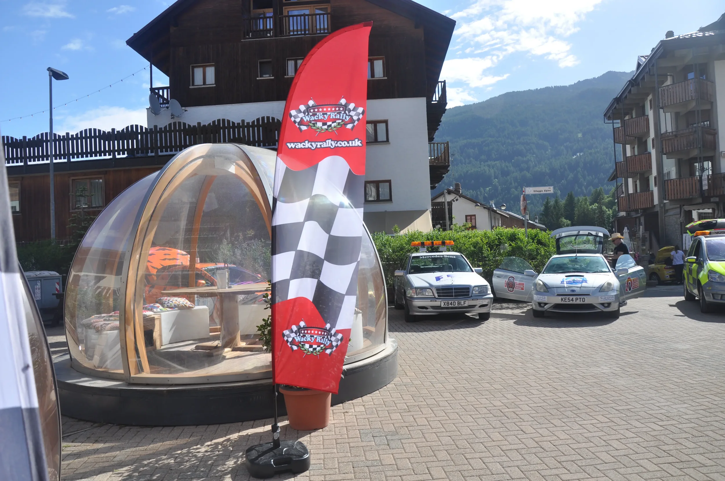 A parking lot with several cars, including police cars, and a stone and glass shelter with a wooden bench inside. There is a red flag with a racing checkered pattern, and the text 'Wacky Rally' and a website address. In the background, there are apar