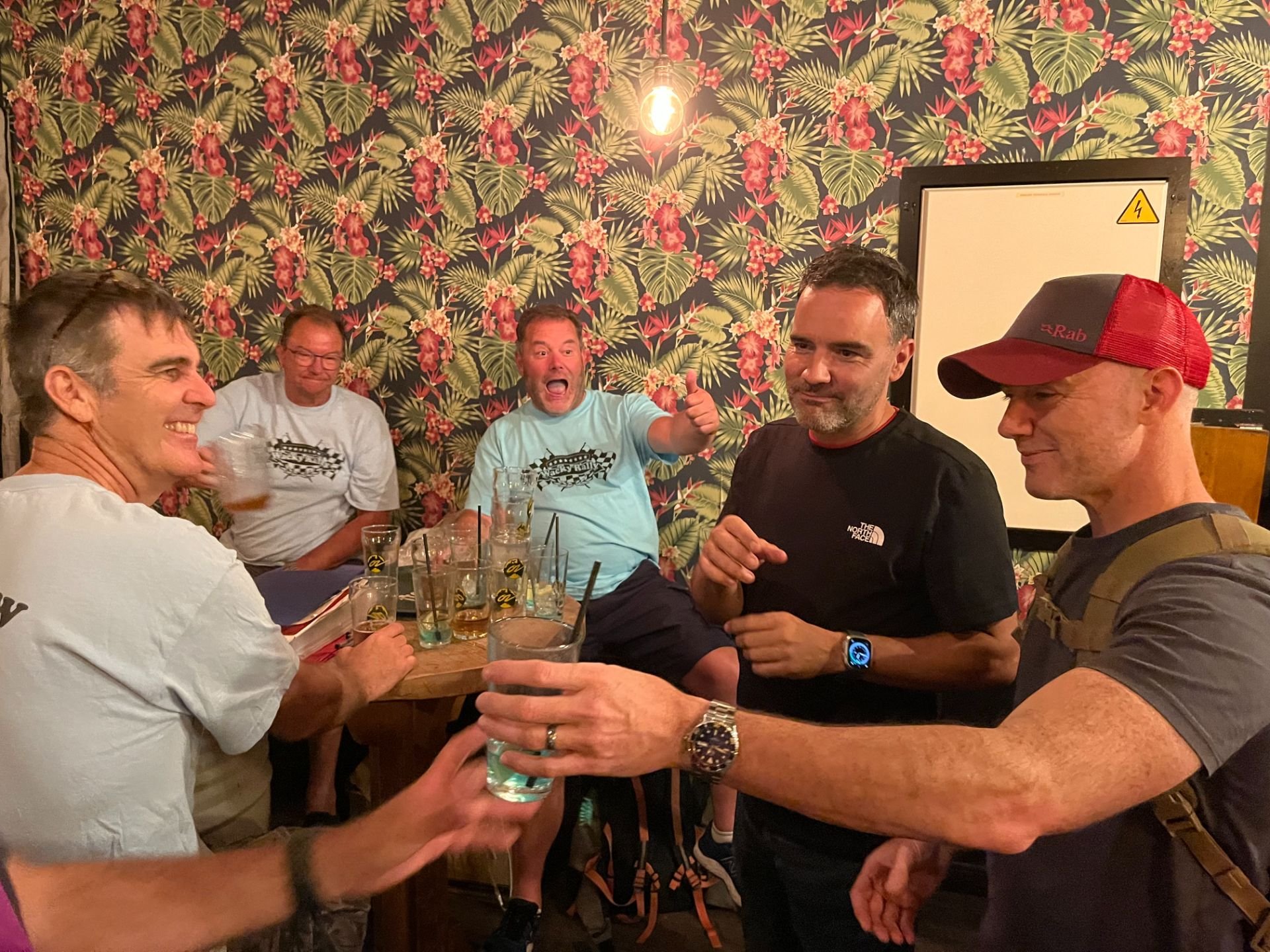 A group of six men socializing at a bar or restaurant, holding drinks, with one man giving a thumbs-up. The background features tropical-patterned wallpaper and a whiteboard, suggesting a casual, lively atmosphere.