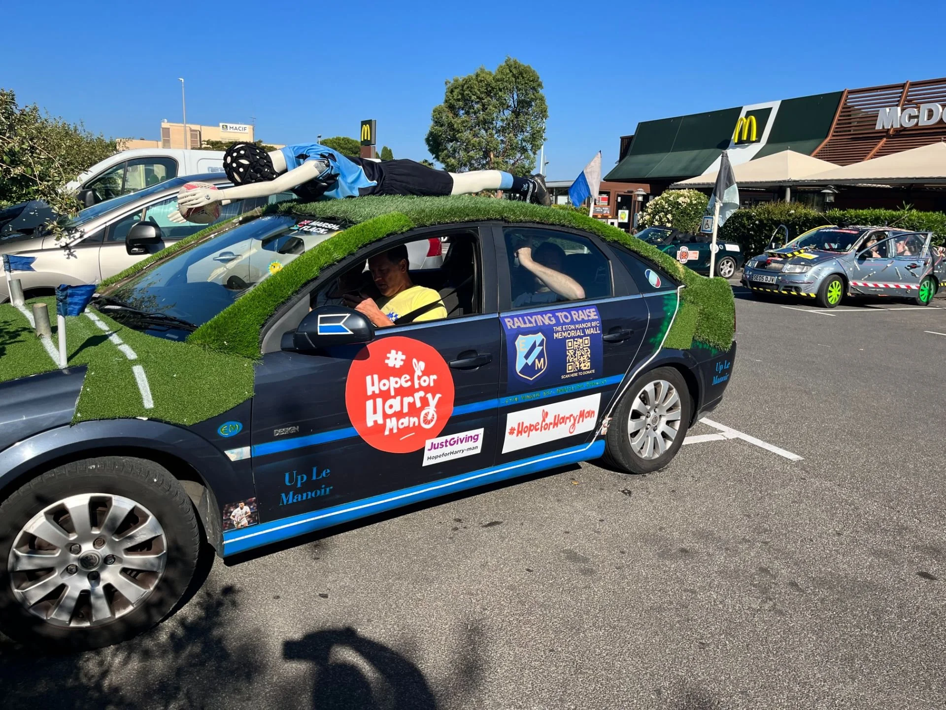 A decorated car with green grass and turf covering parts of it, featuring banners with messages supporting a cause, with a mannequin soccer player on the roof and a person inside the car, parked in a lot near a McDonald's restaurant.
