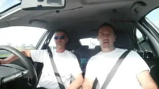 Two men sitting in the front seats of a car, both wearing white shirts and sunglasses, with the driver holding the steering wheel.