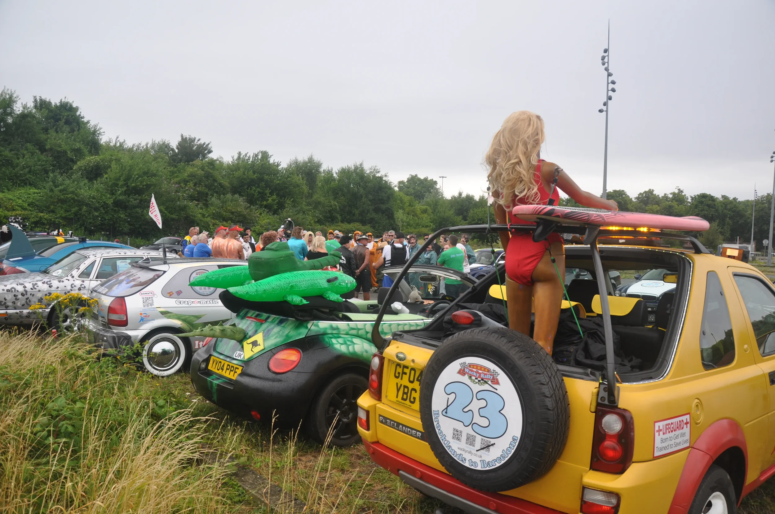 A large gathering of people and parked cars outdoors, with a woman standing in the back of a yellow car holding a pink surfboard, and lots of inflatable floats on the cars.