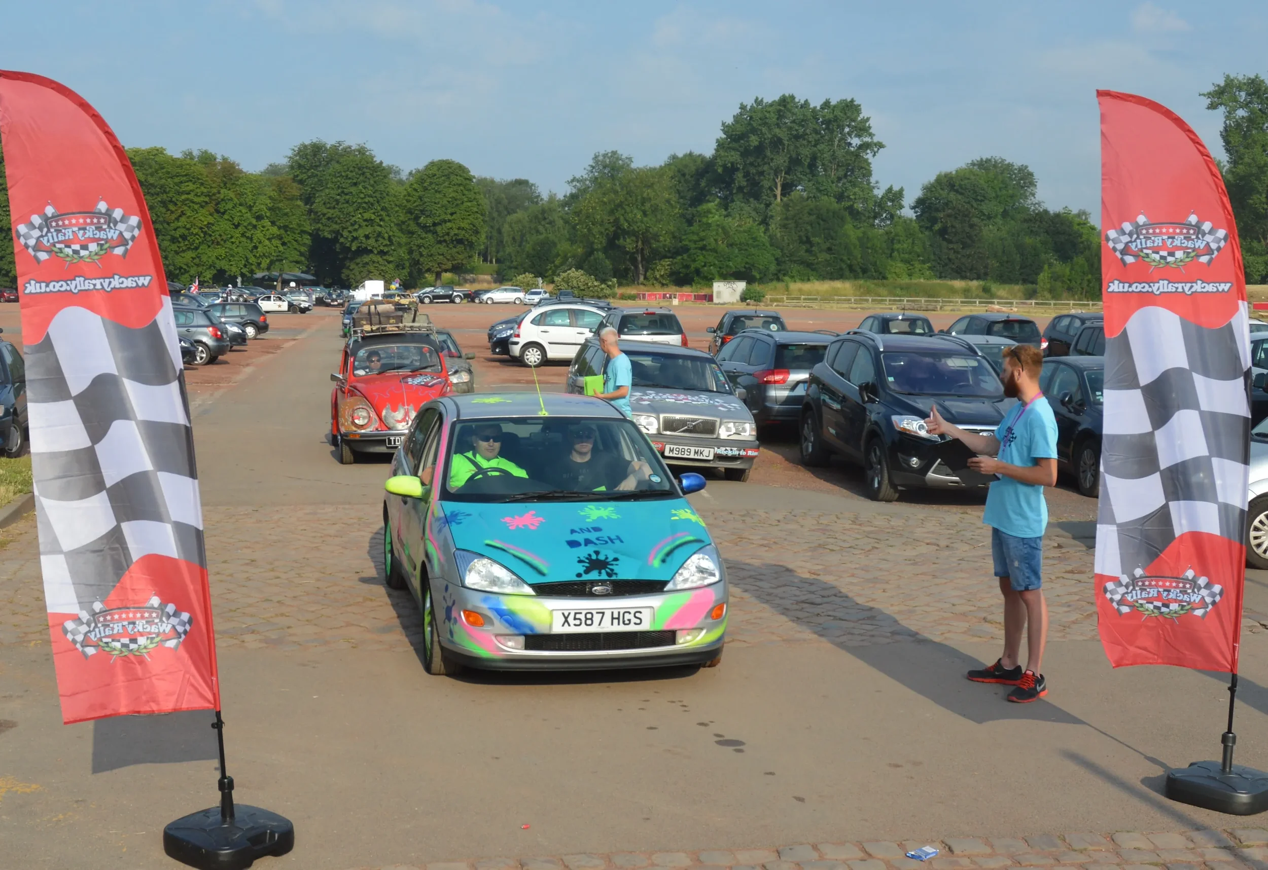 Colorful car participating in a race event at a parking lot with a starting line marked by red flags. A person stands nearby with a clipboard, while two people sit inside the car. In the background, several parked cars and a row of trees are visible 
