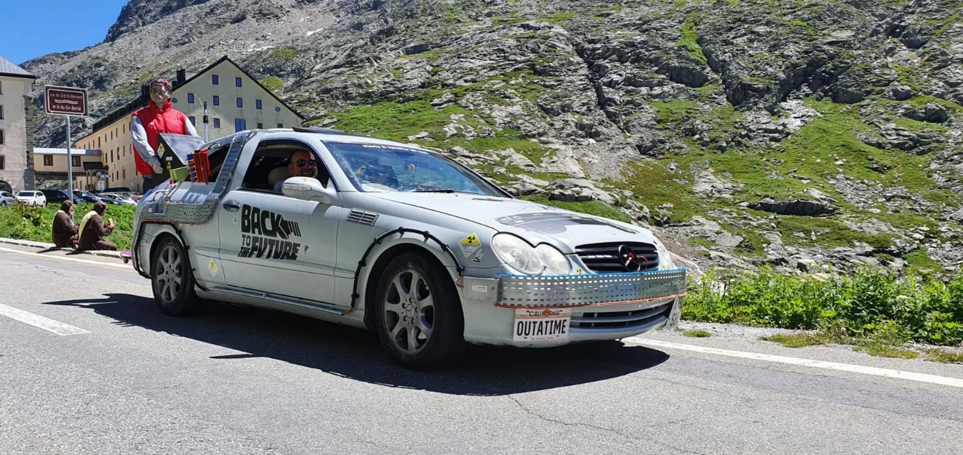 A modified silver Mercedes-Benz car with various decals and additions, including a large metal plate on the front and side, and the phrase 'BACK TO THE FUTURE' on the side. The car has a California license plate reading 'OUTATIME' and is driving in a
