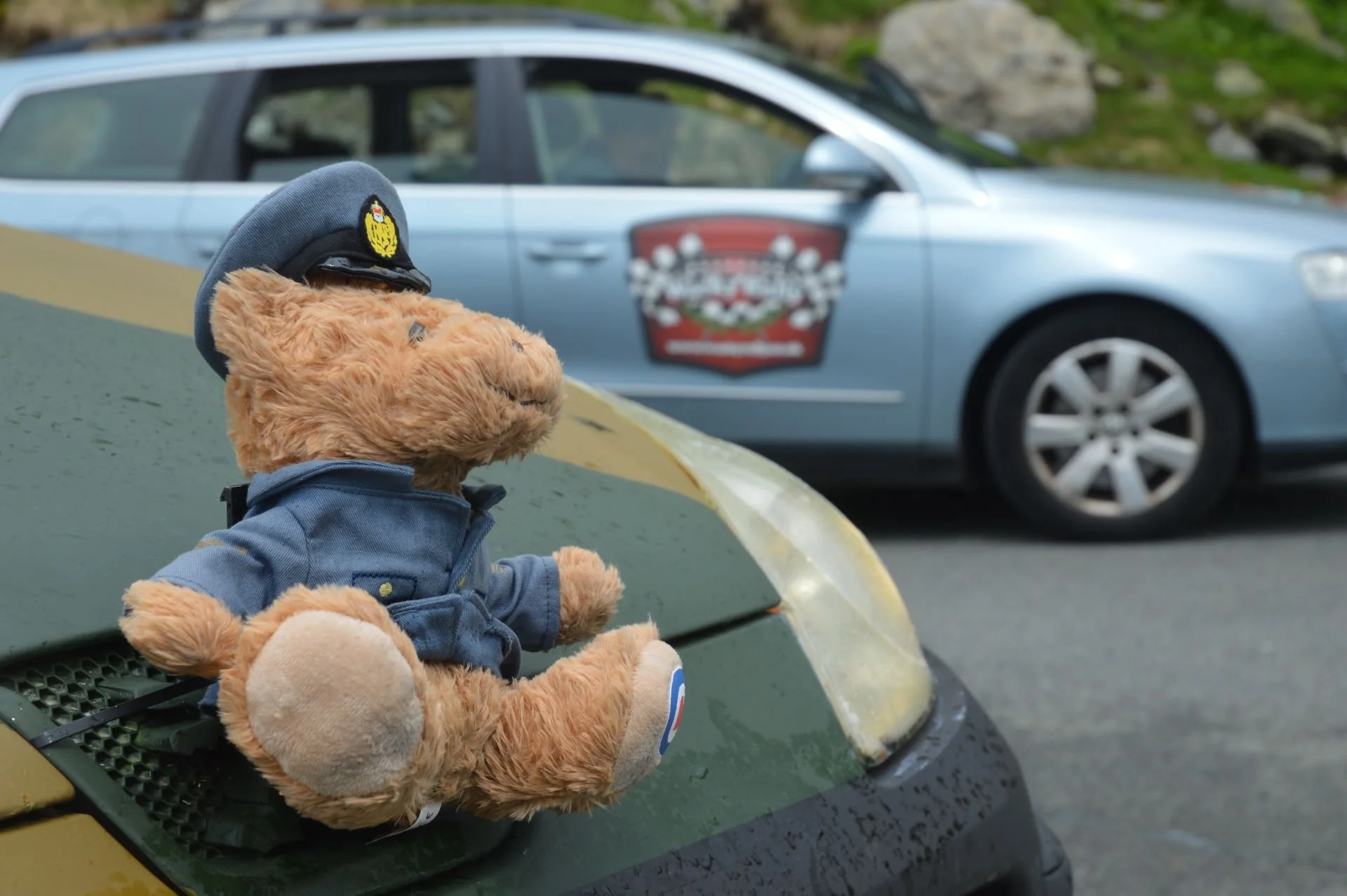Teddy bear wearing a police hat and jacket sitting on the hood of a yellow and black vehicle with a greenish hue, with a light blue police car in the background.