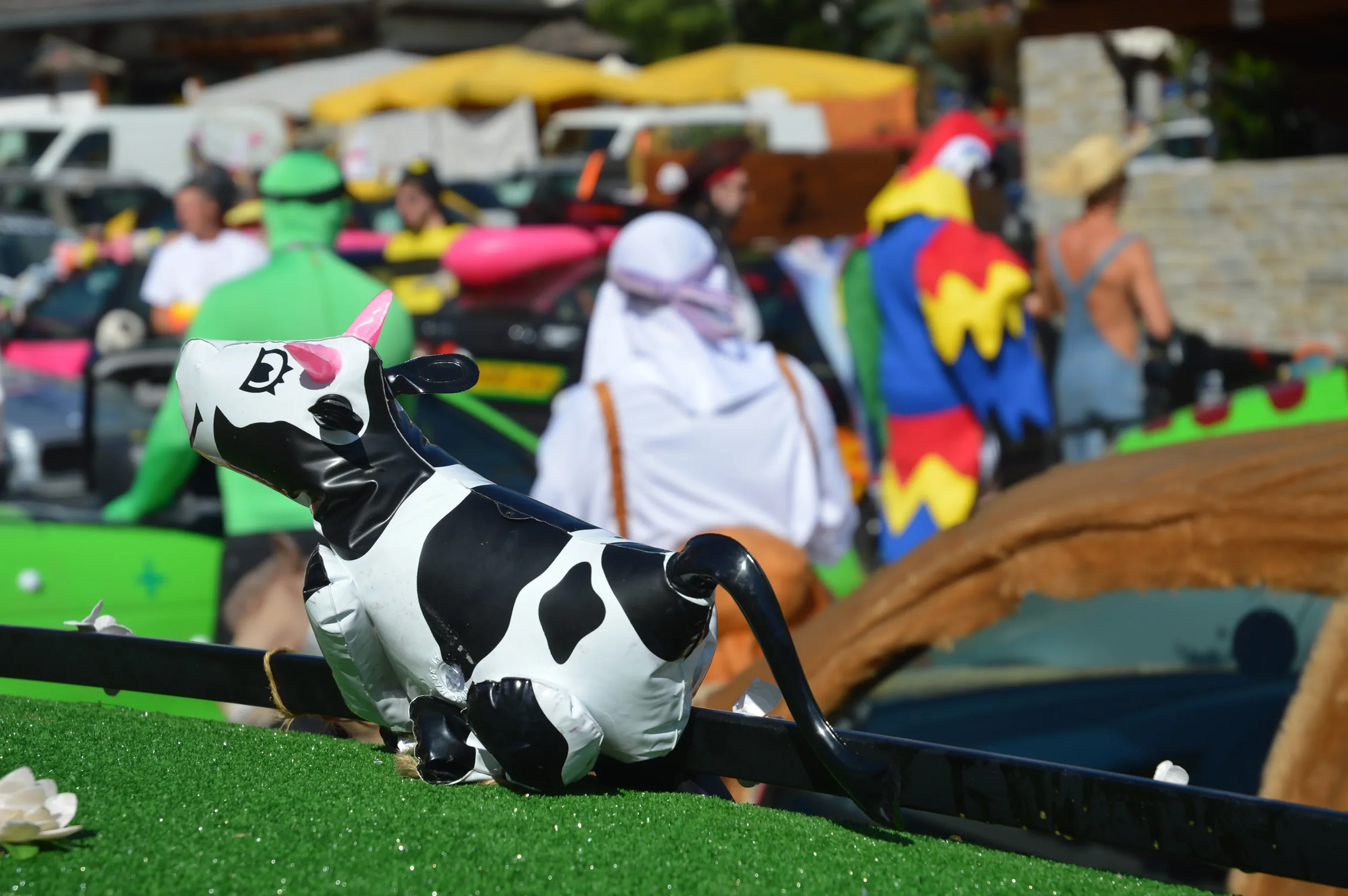 A black and white inflatable cow with pink accents, placed on a green surface in an outdoor setting with people in costumes in the background.