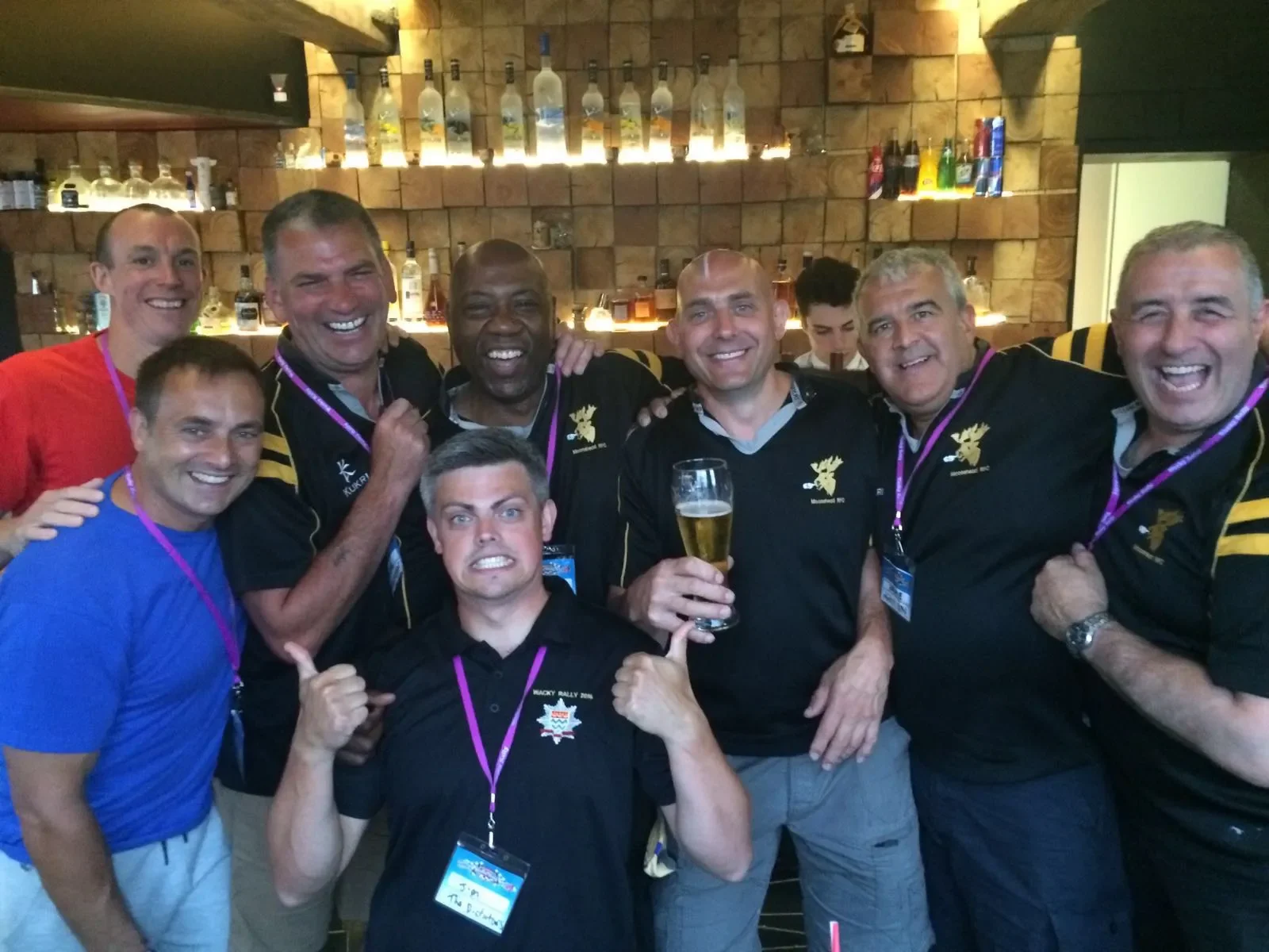 Group of men at a celebration in a bar, smiling and posing for the camera, some wearing team shirts and badges, one holding a beer, in front of a brick wall with bottles on shelves.
