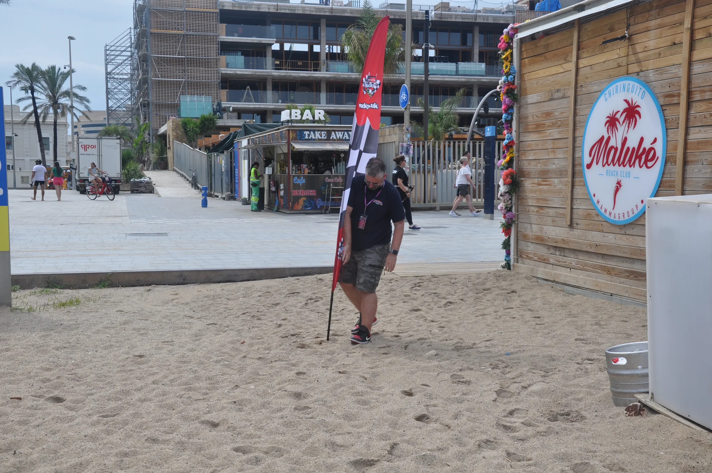 A man in a black shirt and camouflage shorts walking on the sandy beach, holding a flagstick upright in the sand, near a wooden structure with a sign that says 'Maluké Beach Club' decorated with flowers. In the background, there are women walking, a 
